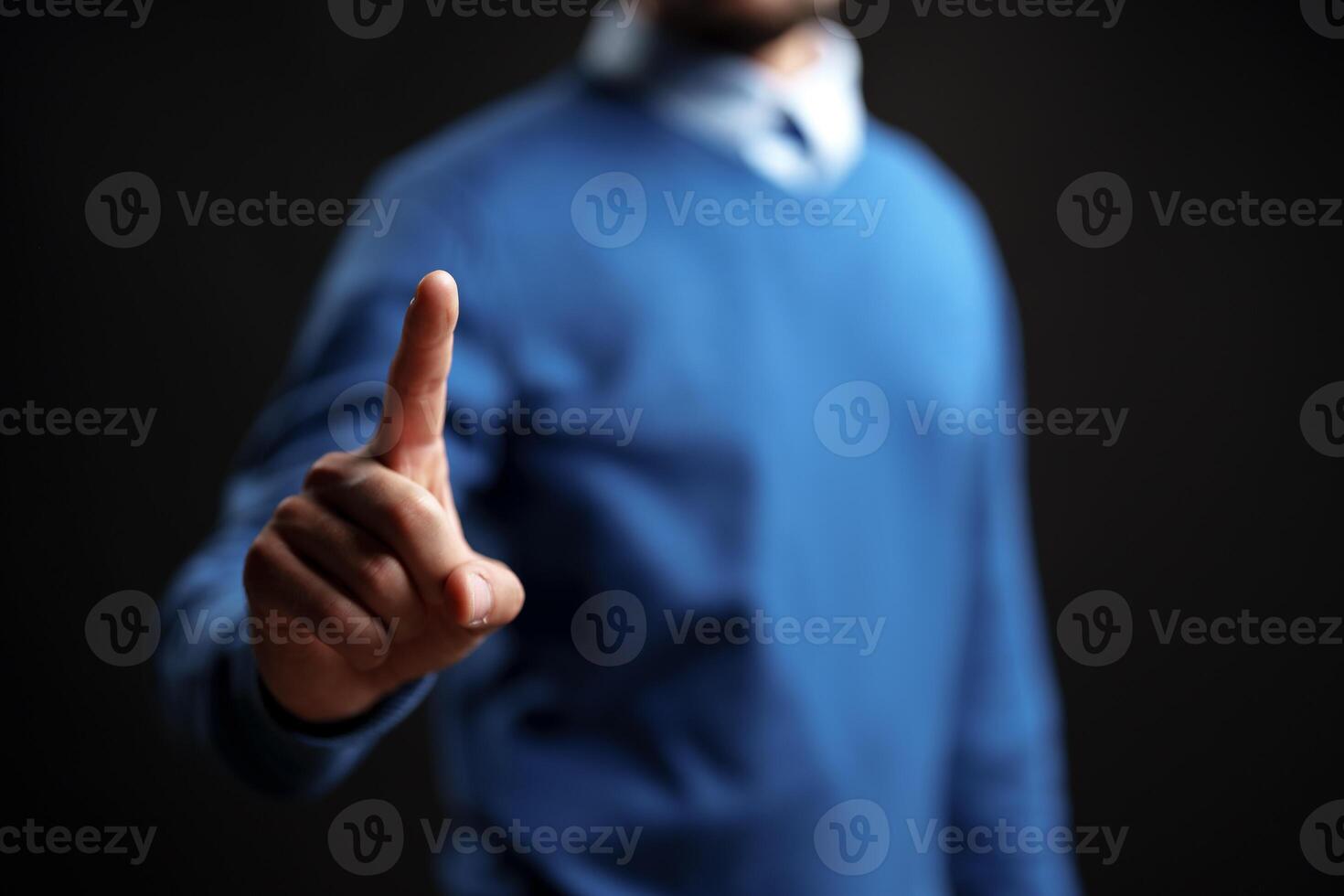 Man signaling to pause with one finger raised, dressed in casual attire against a dark background with soft lighting photo
