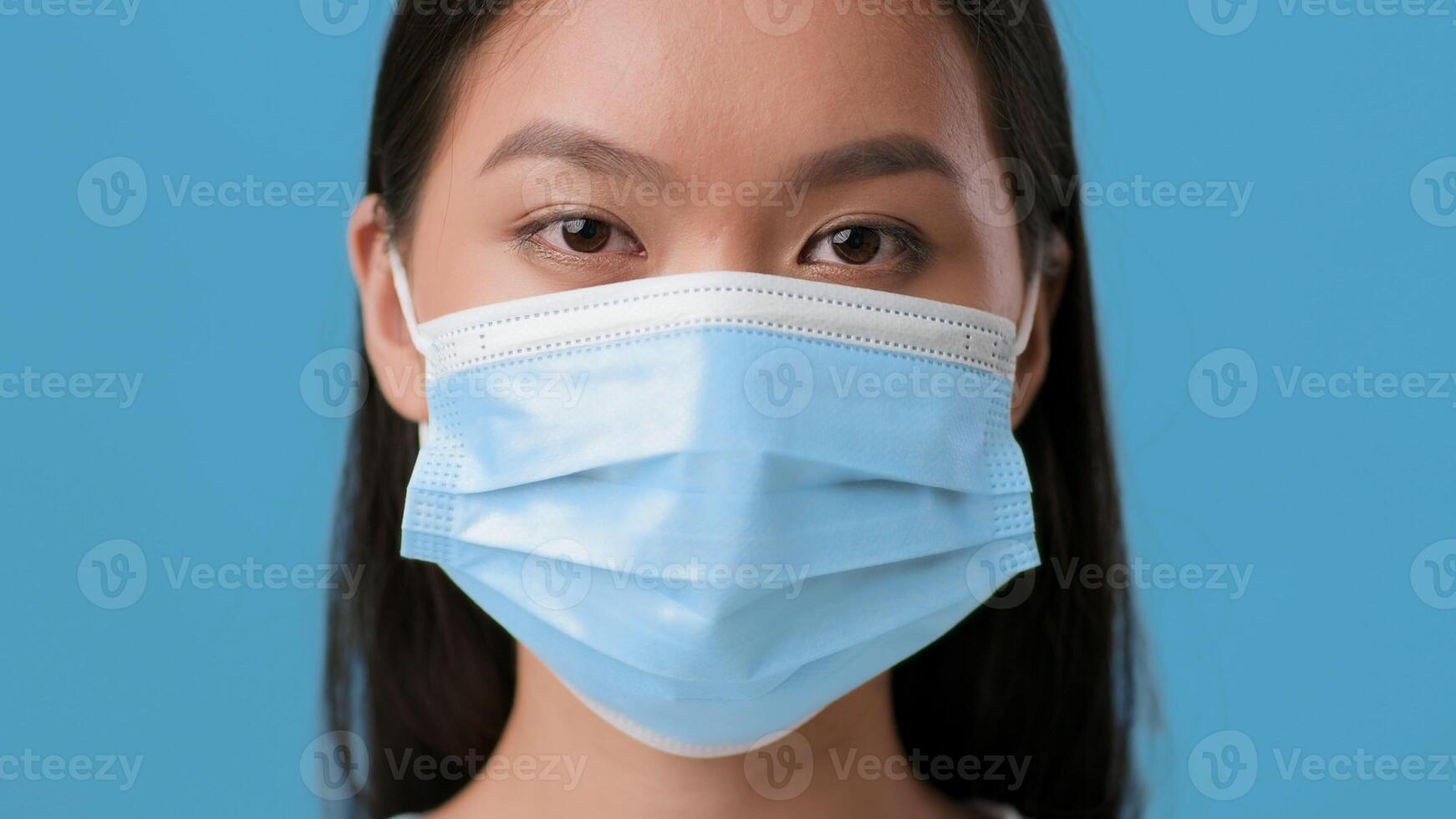 A young woman of Asian descent stands with a neutral expression against a vibrant blue background. She is wearing a medical mask, reflecting the current health climate and lifestyle choices. photo