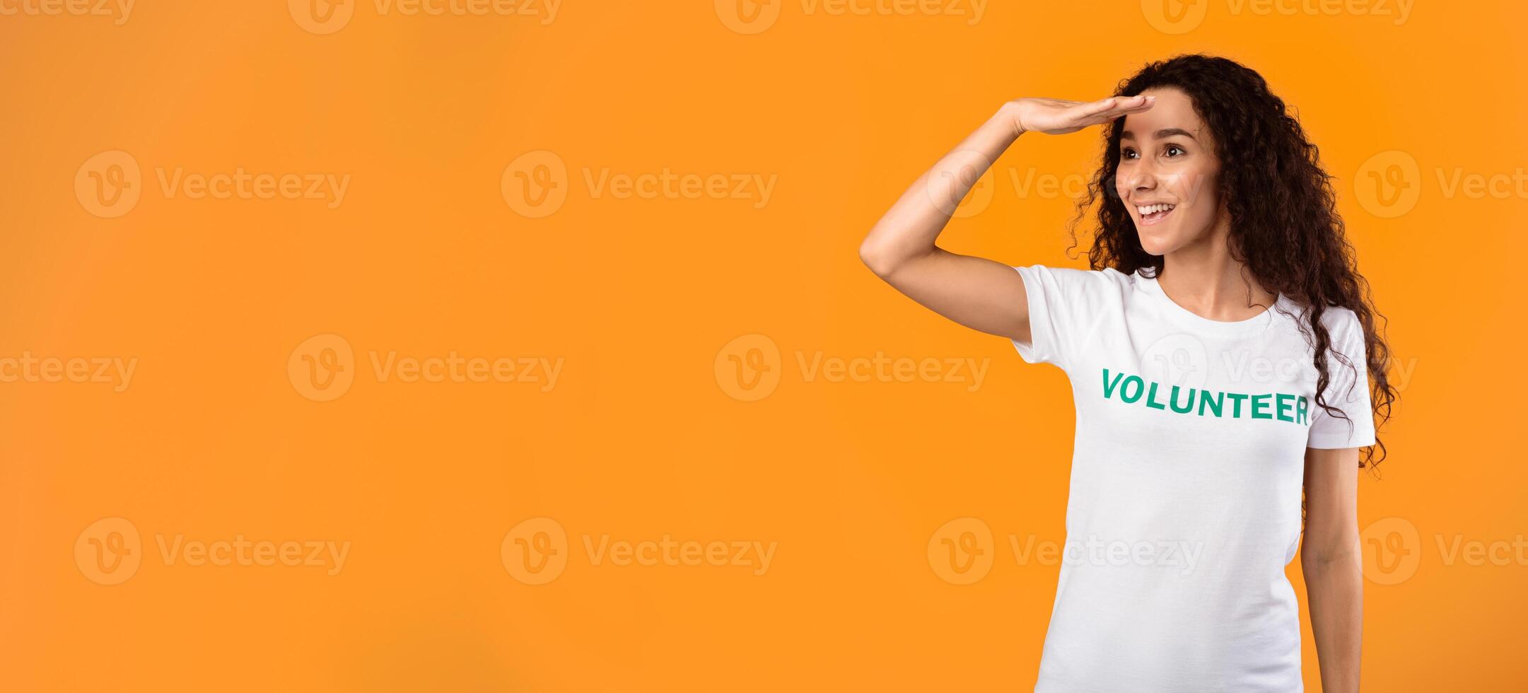 Female Volunteer Looking Aside At Free Space For Advertisement Text Holding Hand Near Eyes Posing Standing Over Yellow Studio Background. Activist Advertising Volunteering Project photo