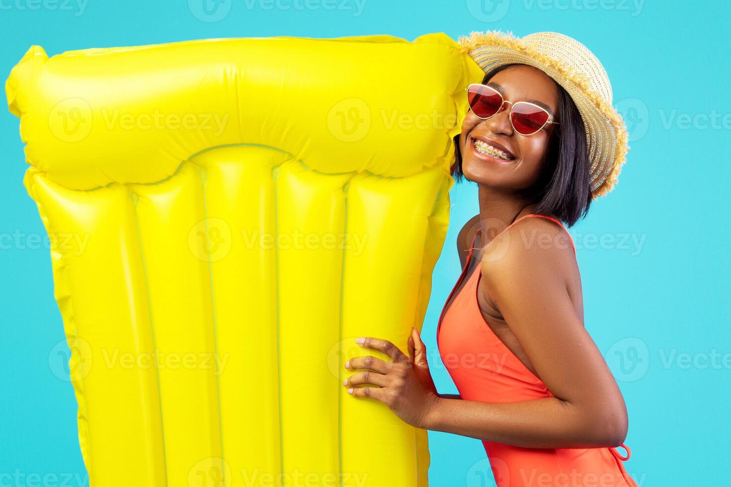 Swimming is fun. Cheerful black woman in swimsuit going to pool with yellow inflatable lilo over blue studio background, full length portrait. African American lady having beach party photo