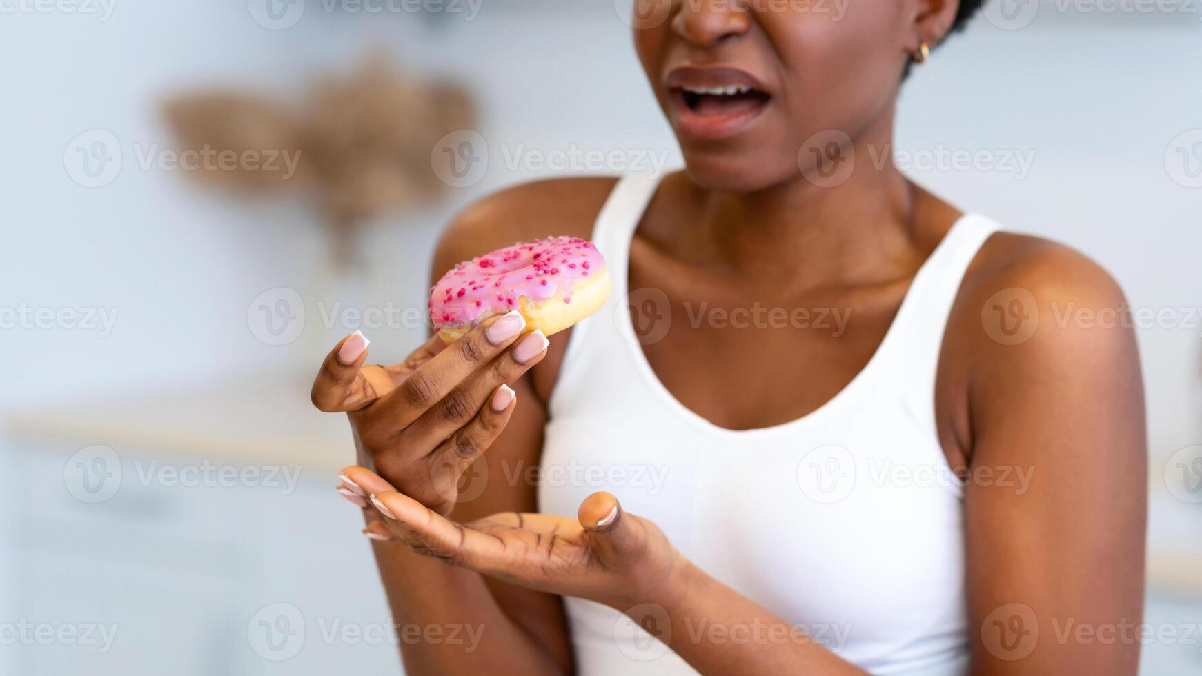Disgusted African American Woman Eating Unhealthy Donut Standing In Modern Kitchen At Home. Bad Nutrition Habit, Junk Food Overeating Concept. Cropped, Selective Focus photo
