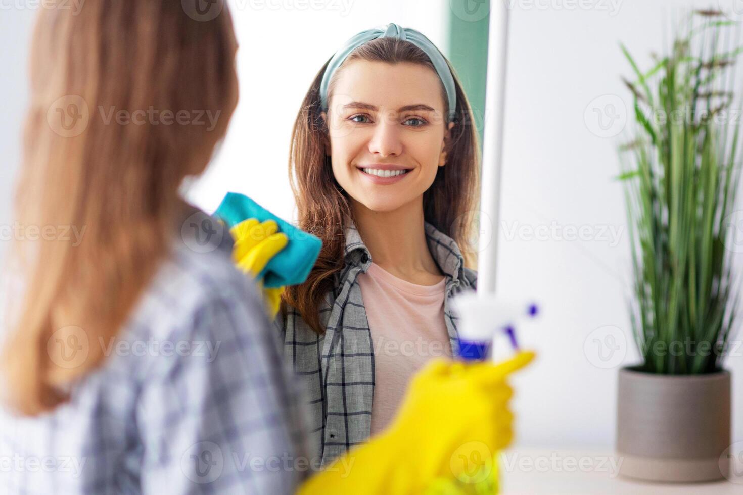 Cheerful millennial housewife wiping mirror with rag, using spray detergent to clean looking glass at home. Young woman doing domestic chores, keeping her apartment tidy photo