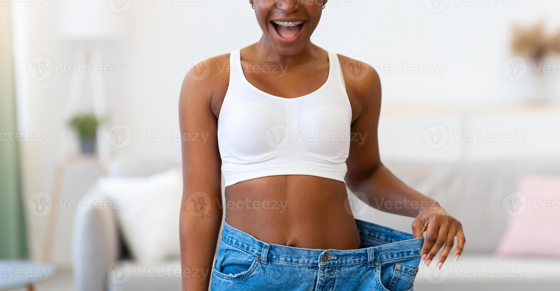 Successful Slimming. Happy Black Woman Wearing Too Big Jeans After Great Weight Loss Standing At Home, Smiling To Camera. Weight-Loss And Dieting Motivation Concept photo