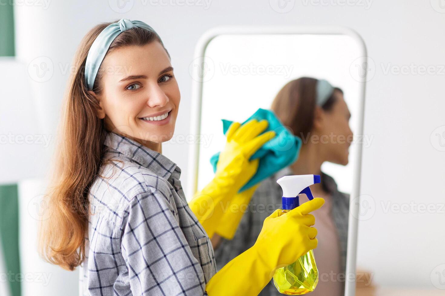 Young Caucasian housewife cleaning mirror, using spray detergent and rag at home. Millennial housemaid doing domestic duties, looking at camera and smiling. Sanitary service concept photo