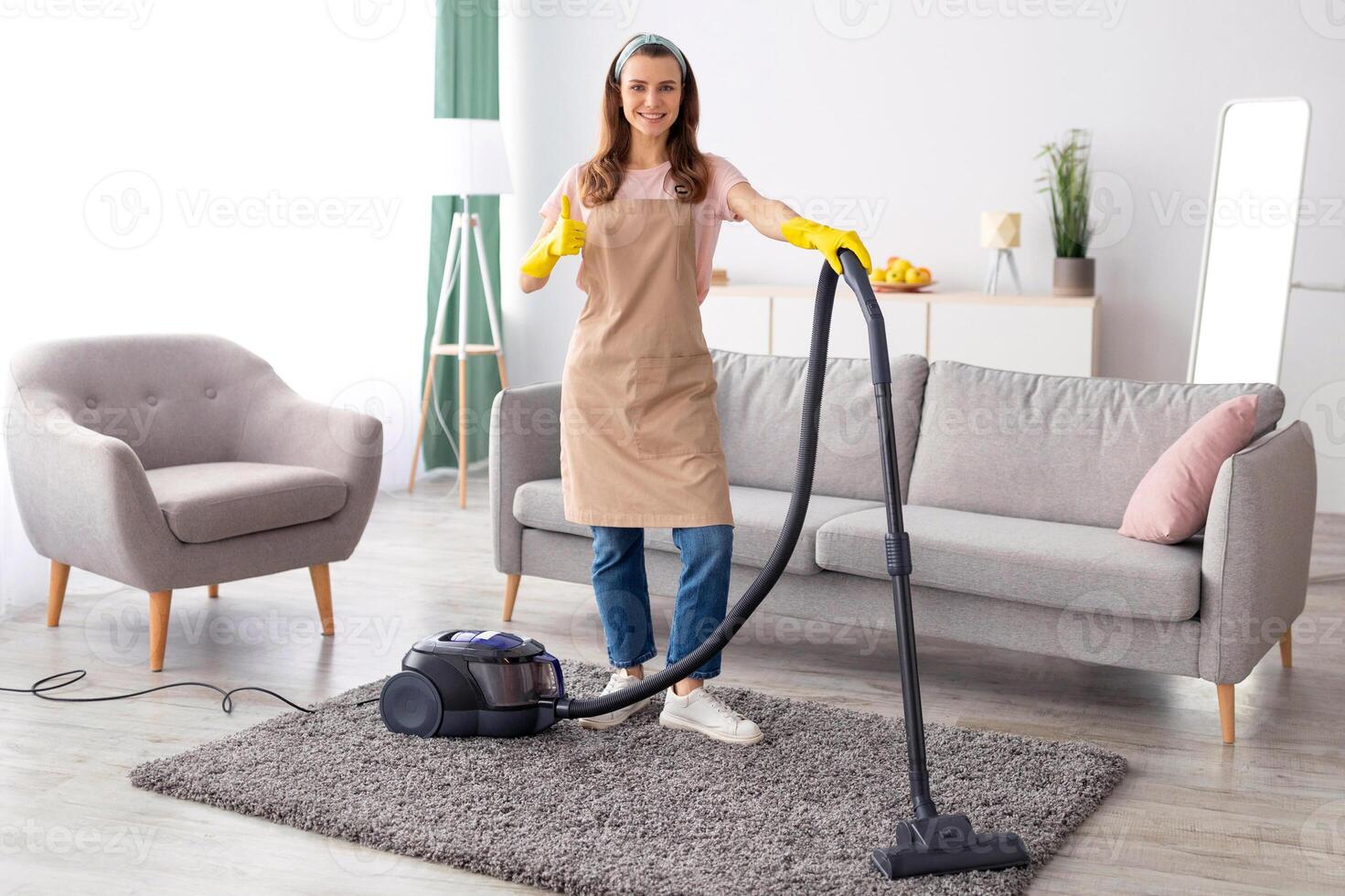 Full length portrait of female cleaning service specialist showing thumb up gesture, standing with vacuum cleaner at tidy house. Young woman in apron and rubber gloves doing domestic chores photo