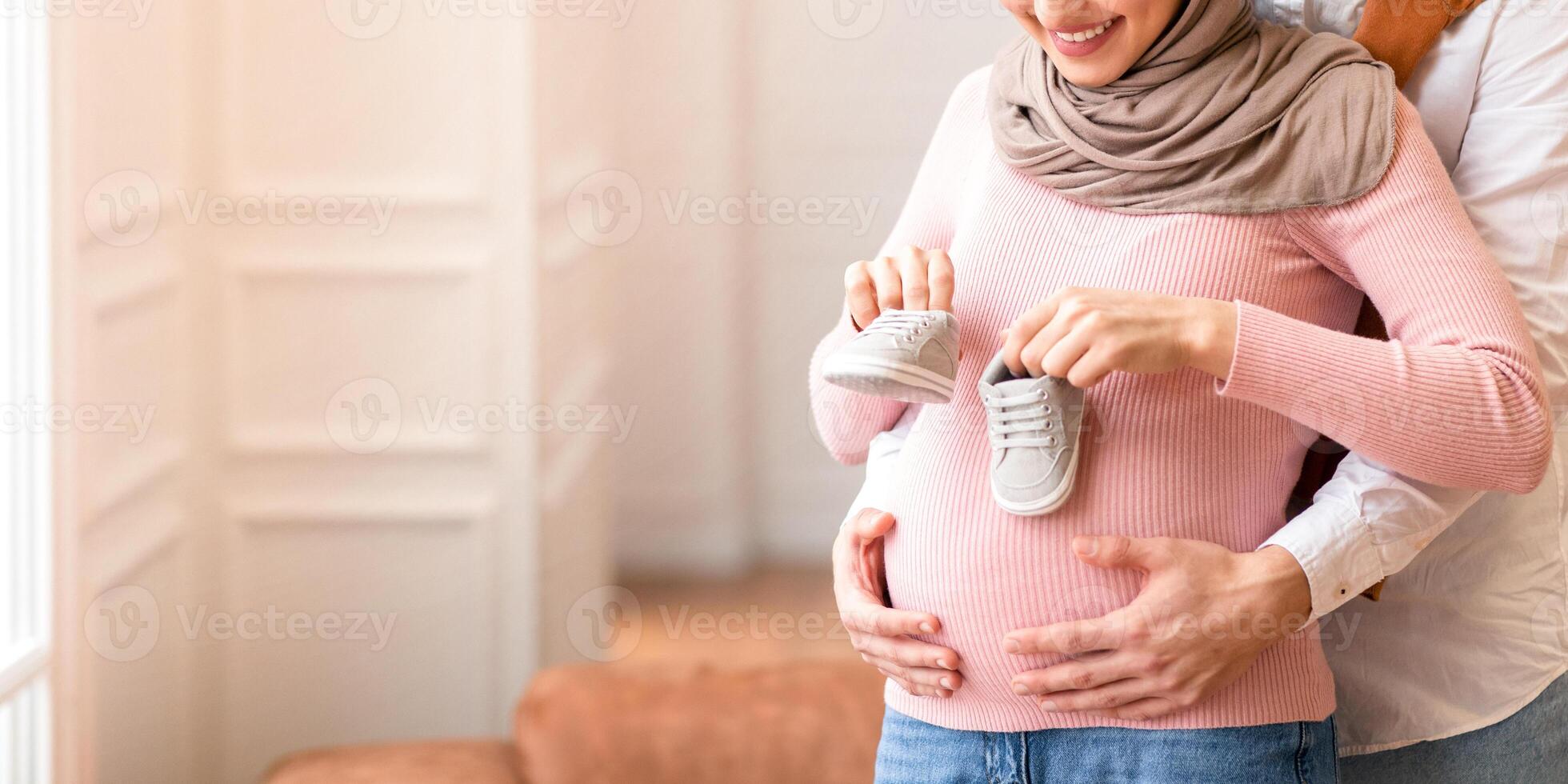 Muslim Husband Holding Tiny Shoes Hugging Pregnant Wife's Belly Hugging Standing At Home. Family Happiness And New Life. Happy Parents-To-Be Awaiting Baby Concept. Free Space For Text photo