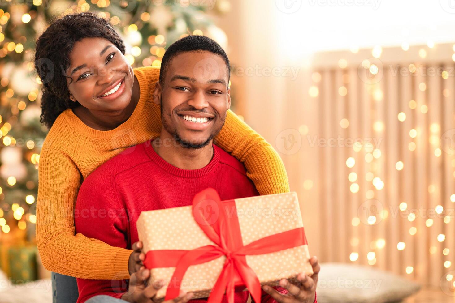 Winter mood, romance and gifts during covid-19 pandemic. Happy millennial african american couple hug, hold gift box with red ribbon in living room interior with Christmas tree and glowing garlands photo