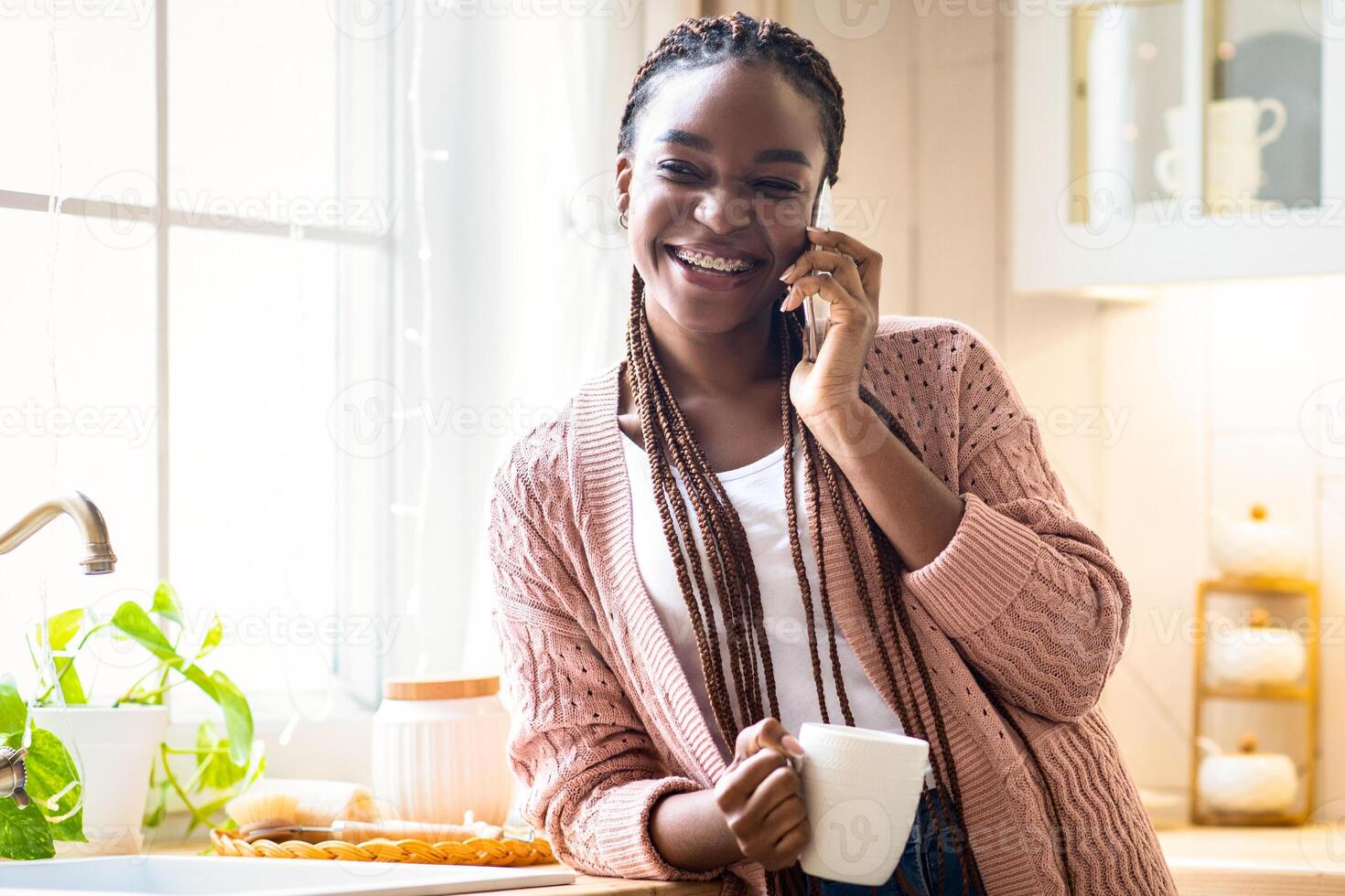 Domestic Leisure. Cheerful African American Lady Drinking Coffee And Talking On Cellphone At Home, Black Woman Having Mobile Conversation And Enjoying Hot Drink In Kitchen Interior, Free Space photo