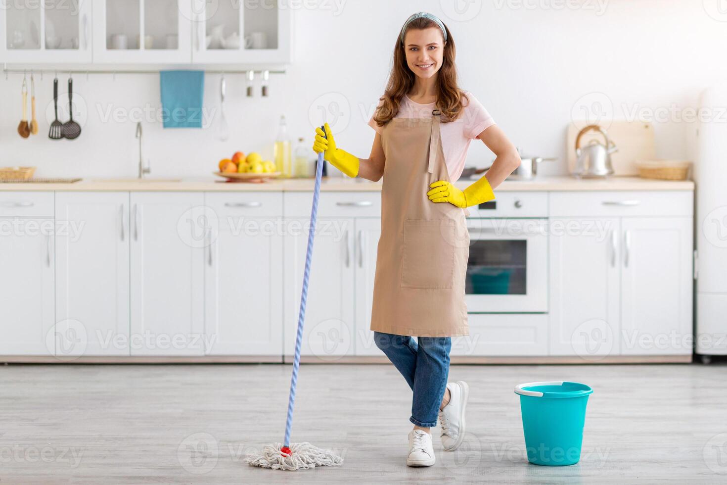 Full length portrait of happy young lady with mop washing floor in kitchen, empty space. Smiling millennial maid or housewife keeping her apartment tidy and clean, doing disinfection photo