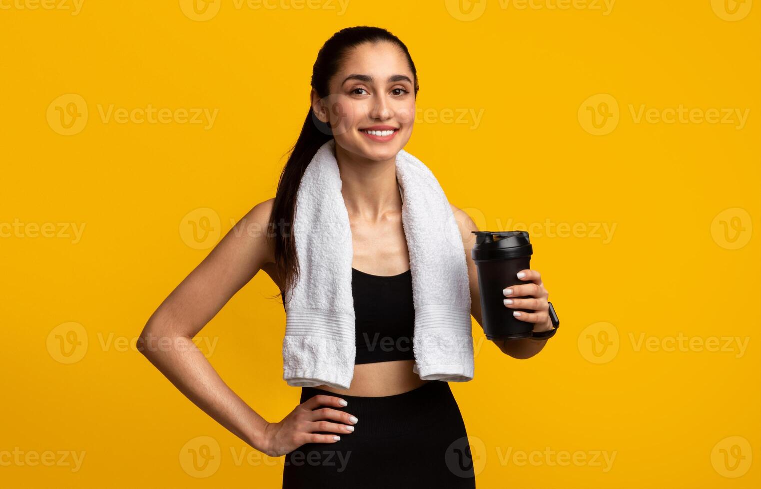 Portrait of sporty beautiful smiling woman in black sportswear and white towel on neck holding shaker with healthy drink, whey protein, fresh water or coffee isolated over orange studio background photo