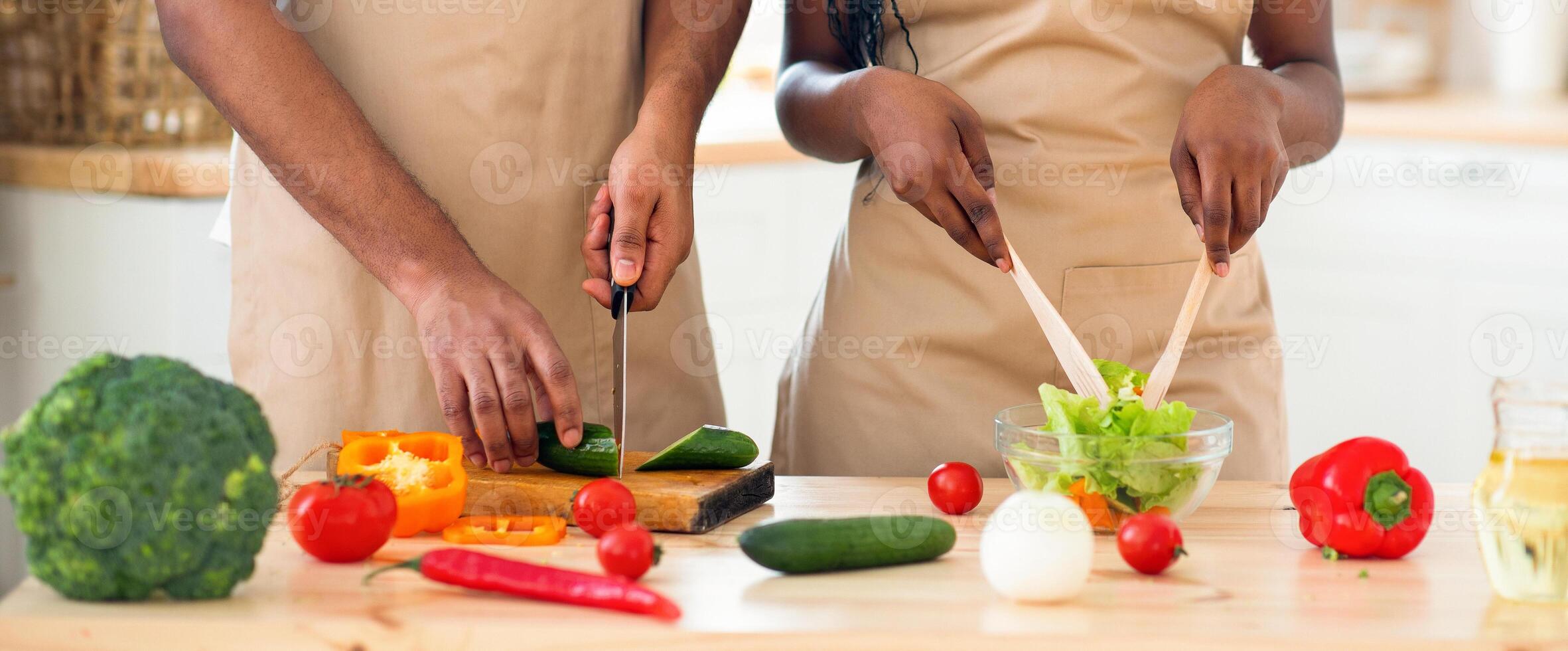 Unrecognizable black couple in aprons preparing vegetable salad together in kitchen. Young african american family cooking healthy dinner together, enjoying making healthy food at come, cropped image photo