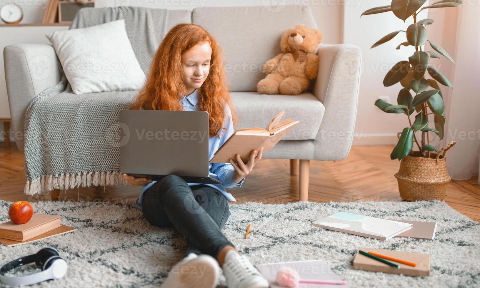 Preparing For Exam. Smart smiling casual gen z girl sitting on the floor carpet at home in living room, leaning on couch, holding paper book and using laptop computer, doing homework photo