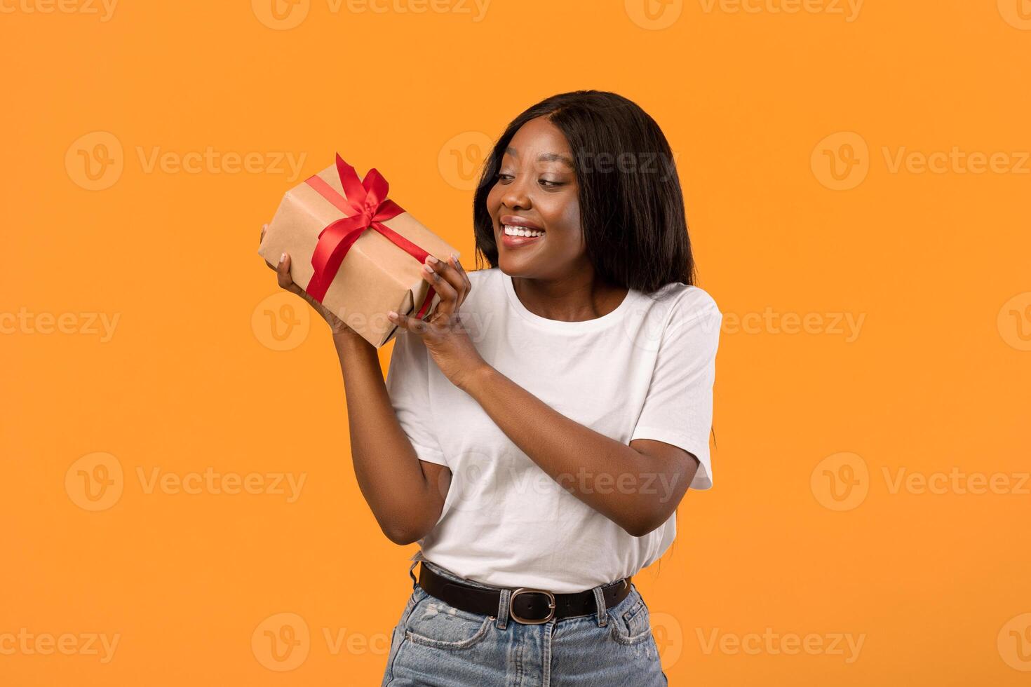 Happy african american young woman holding gift box next to her ear, wondering what is inside, orange studio background, copy space. St. Valentine Day, Birthday, gift, present, surprise concept photo