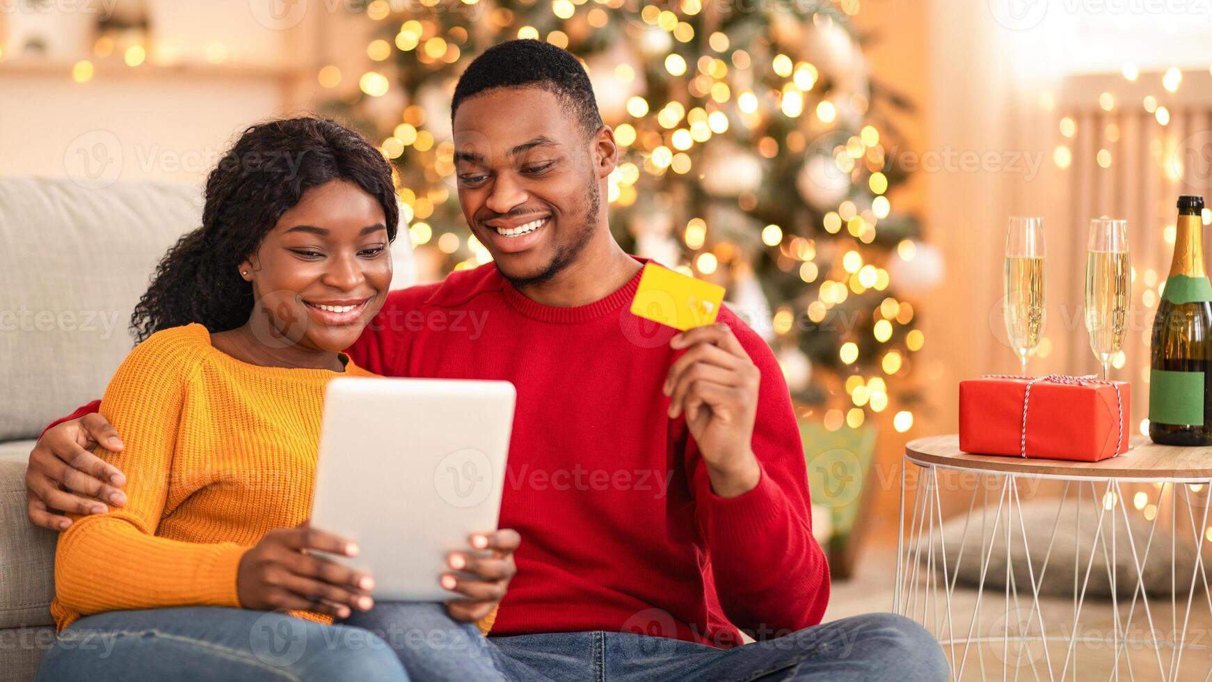 Holiday sale and huge discounts for online shopping. Happy millennial african american guy and woman use tablet and show credit card in cozy living room interior with Xmas tree and glowing garlands photo
