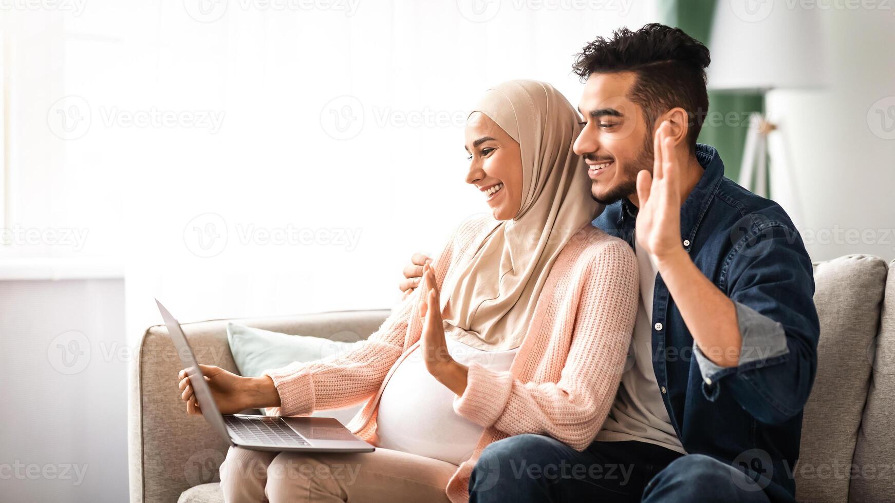 Cheerful Pregnant Muslim Couple Using Laptop At Home For Call, Happy Expectant Middle Eastern Spouses Talking To Family Or Friends Via Web Conference, Waving Hands At Computer Camera, Side View photo
