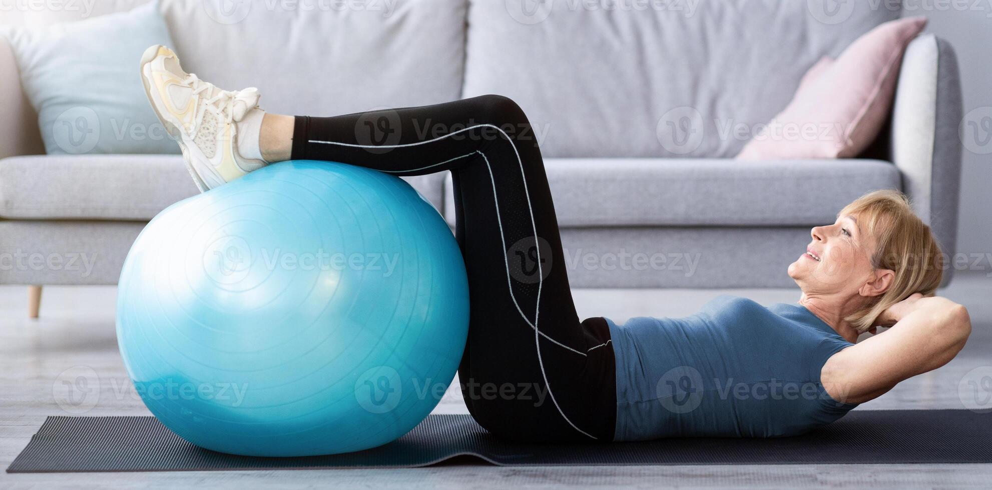 Fit mature lady exercising her abs muscles with fitball on yoga mat at home, copy space. Side view of sporty senior woman leading active lifestyle, working out during coronavirus lockdown photo