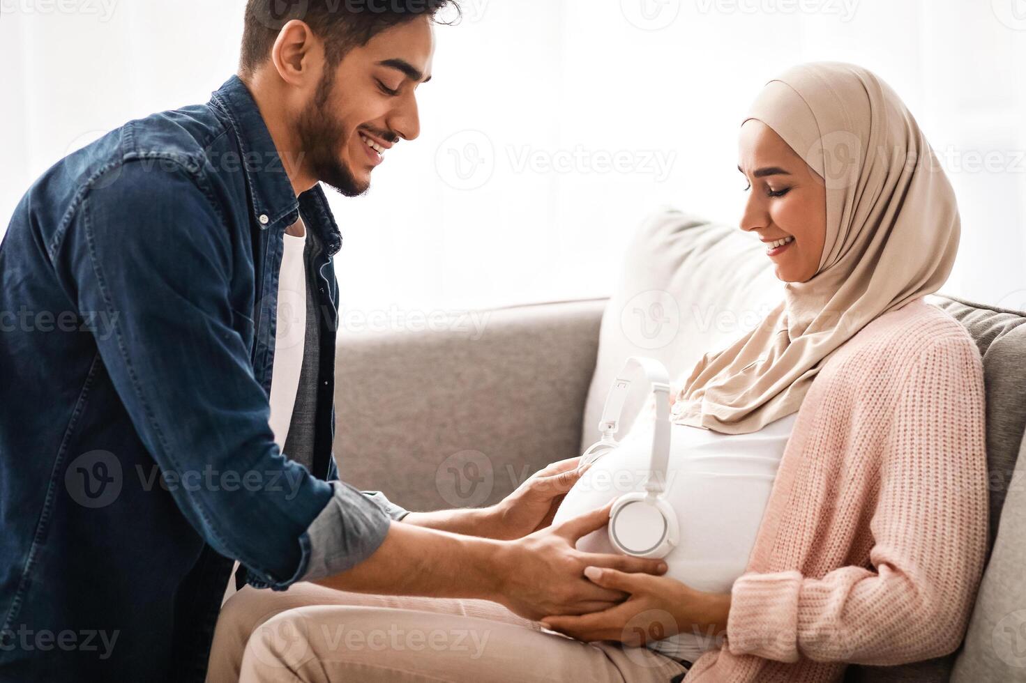 Music For Baby In Womb. Loving Arab Husband Placing Wireless Headphones On Pregnant Wife's Belly While They Resting Together On Couch In Living Room At Home, Muslim Spouses Enjoying Future Parenthood photo