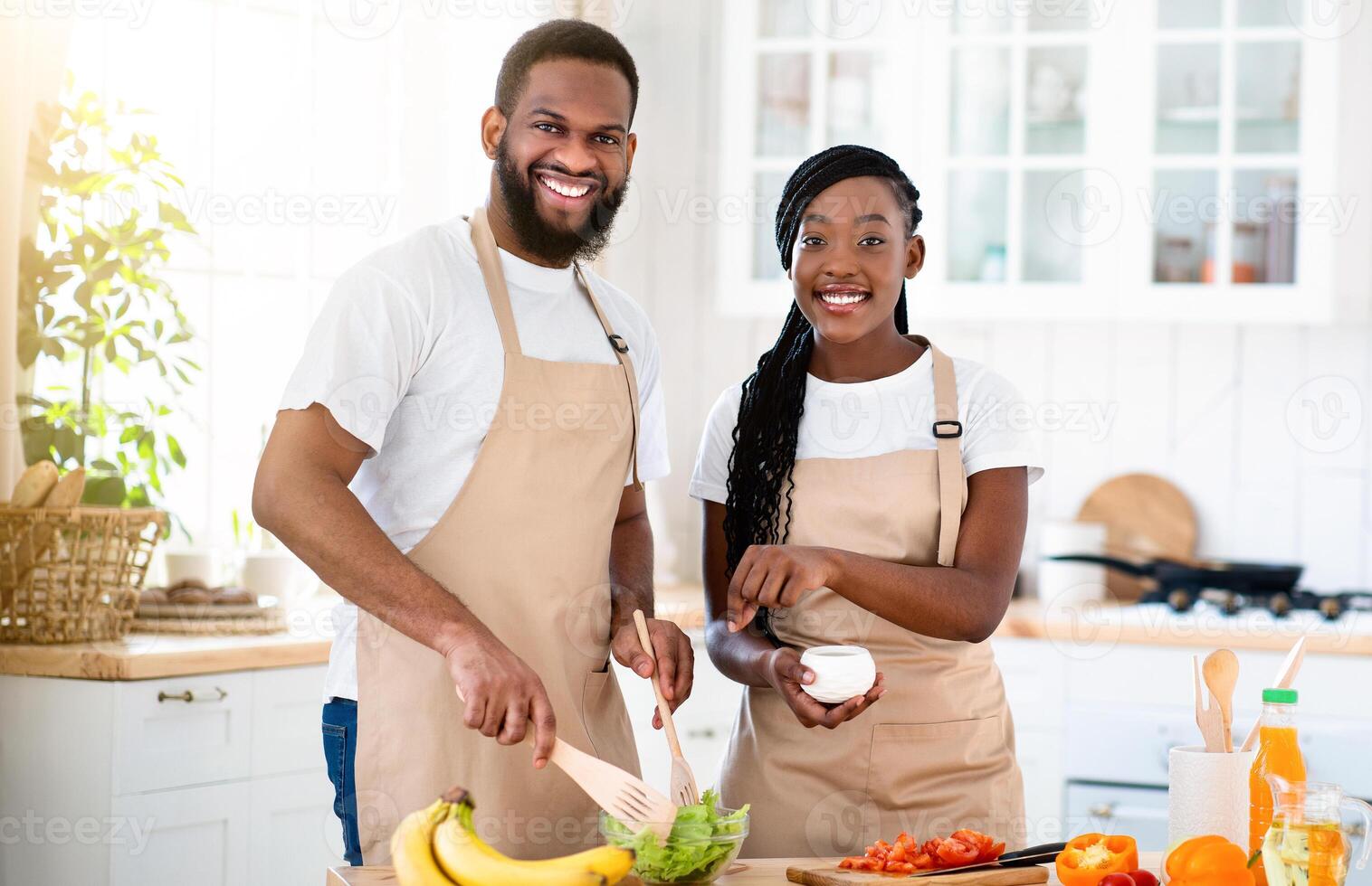 Happy Millennial African American Couple Wearing Aprons Cooking Healthy Lunch Together In Kitchen, Cheerful Black Spouses Enjoying Preparing Food At Home, Looking At Camera And Smiling, Free Space photo