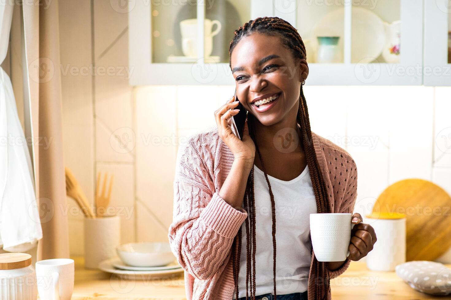 Cheerful black woman talking on mobile phone and drinking coffee in kitchen at home, positive black lady holding mug with hot drink and enjoying pleasant conversation with friend, free space photo