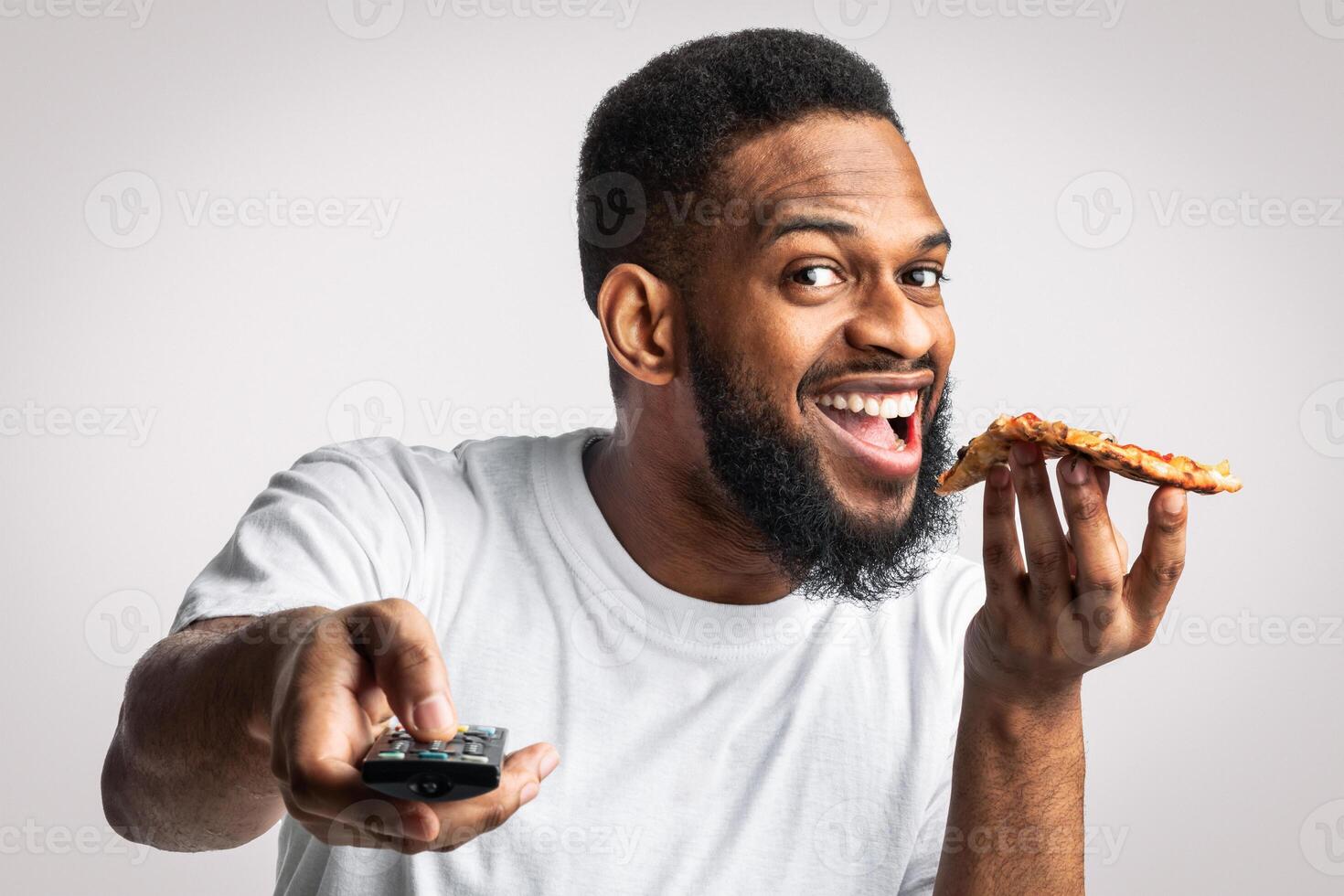 Joyful African Millennial Man Eating Pizza Watching TV Pointing Remote Control At Camera Posing In Studio Over White Background. Selective Focus. Lazy Lifestyle And Unhealthy Nutrition Habits. photo