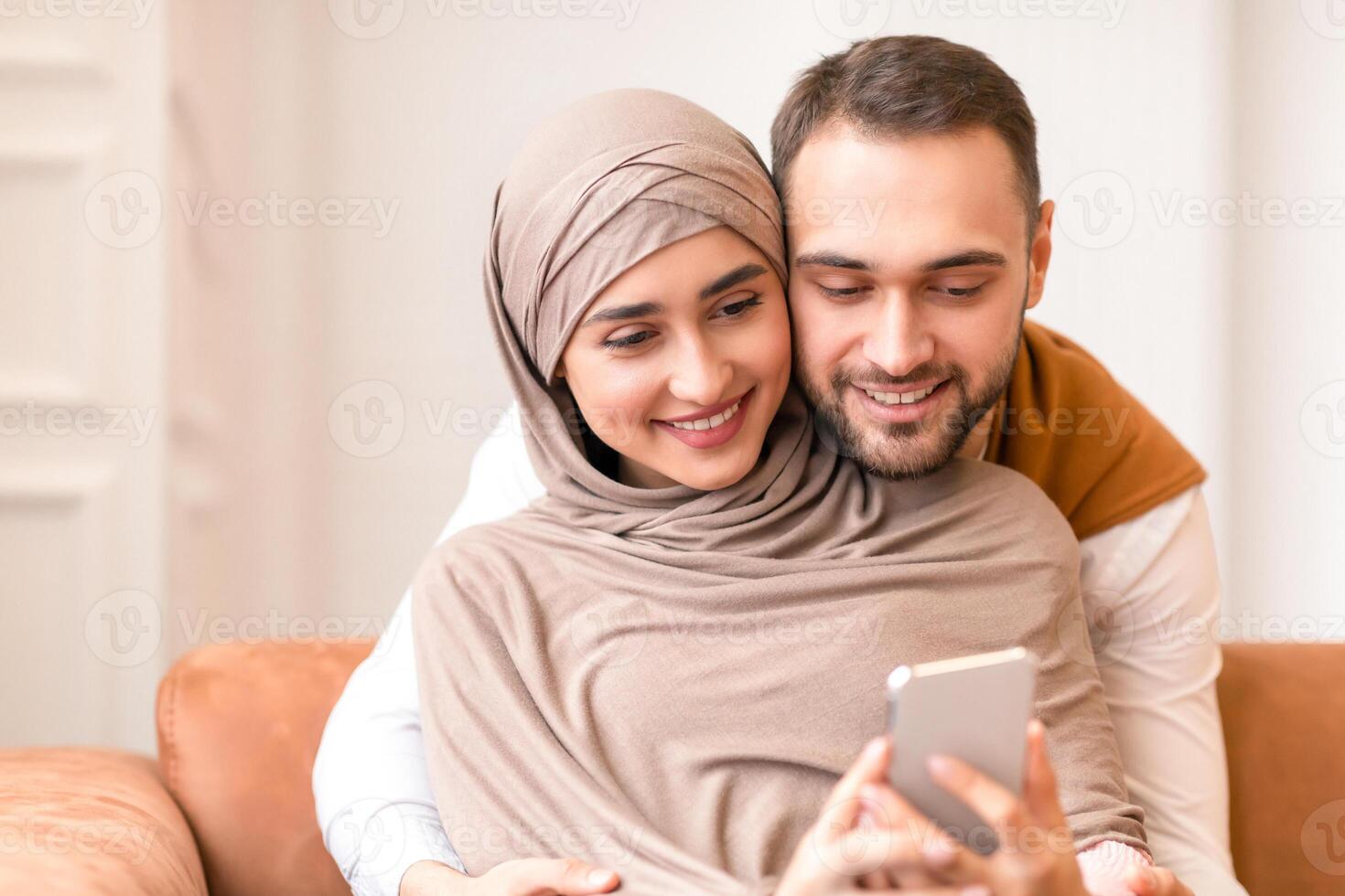 New Application. Happy Muslim Couple Using Mobile Phone Together Hugging Sitting On Couch At Home. Young Arab Spouses Browsing Internet On Cellphone Indoors. Modern Technology And Communication photo