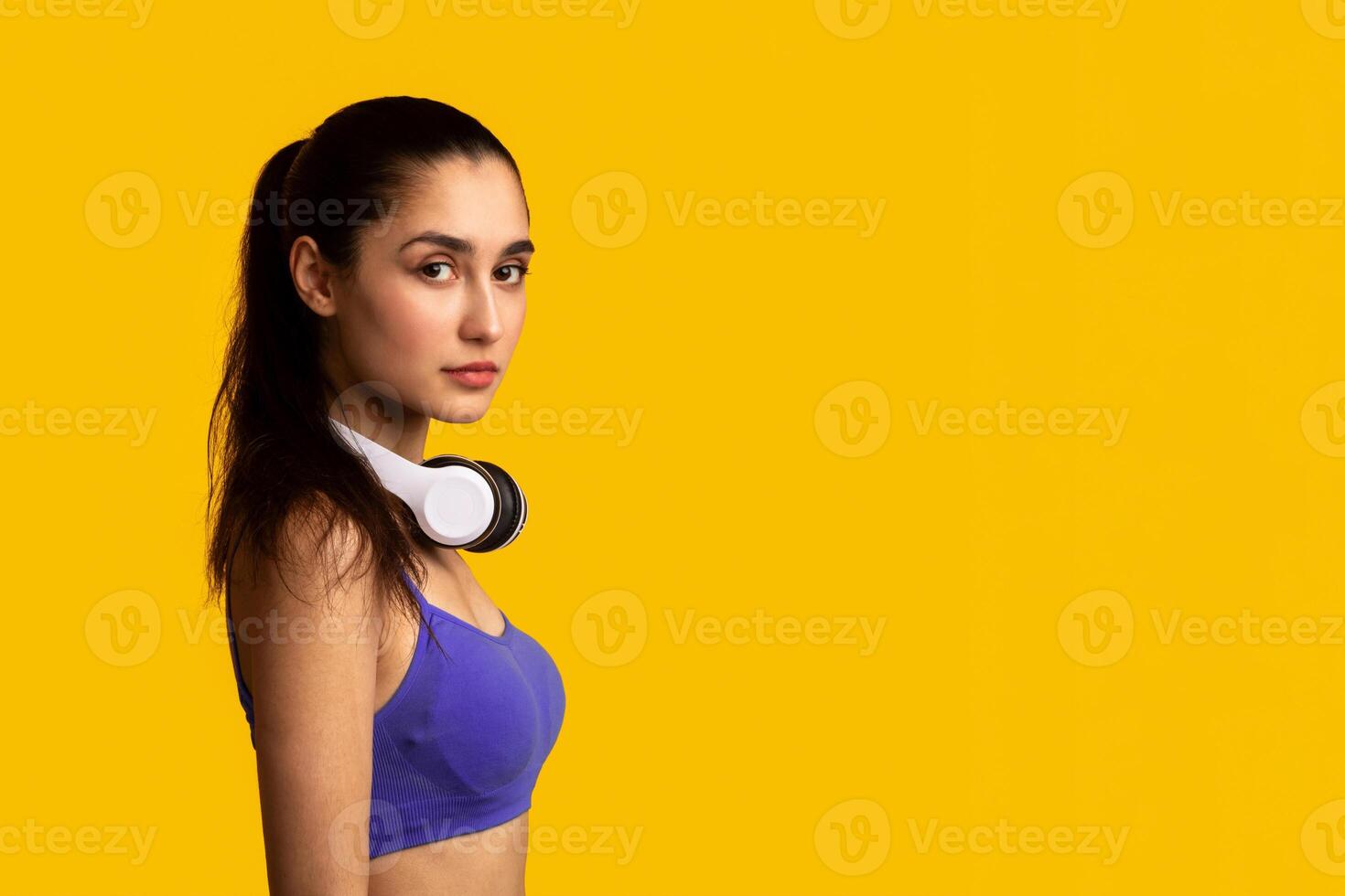 Healthy Lifestyle, Workout With Music. Portrait Of Half-Turned Determined Young Lady With Ponytail Wearing Wireless Headphones On Neck Looking At Camera Isolated Over Orange Studio Background photo