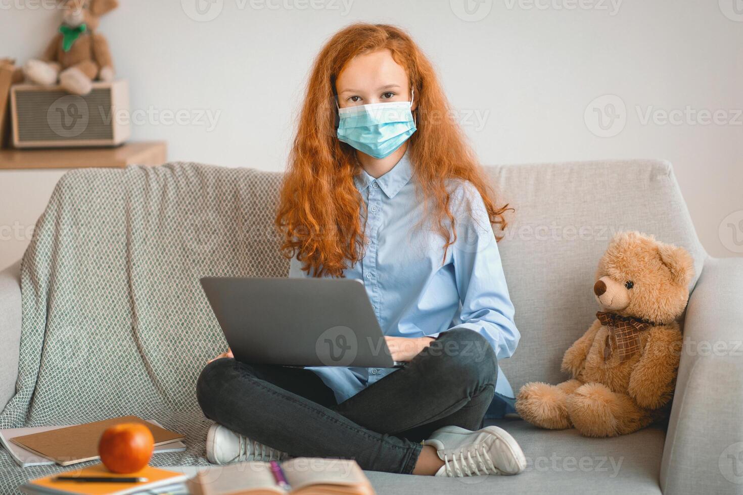 Remote Education During Quarantine, Lockdown. Portrait of girl wearing medical disposable face mask studying at home on laptop computer, sitting on couch and looking at camera, doing homework photo