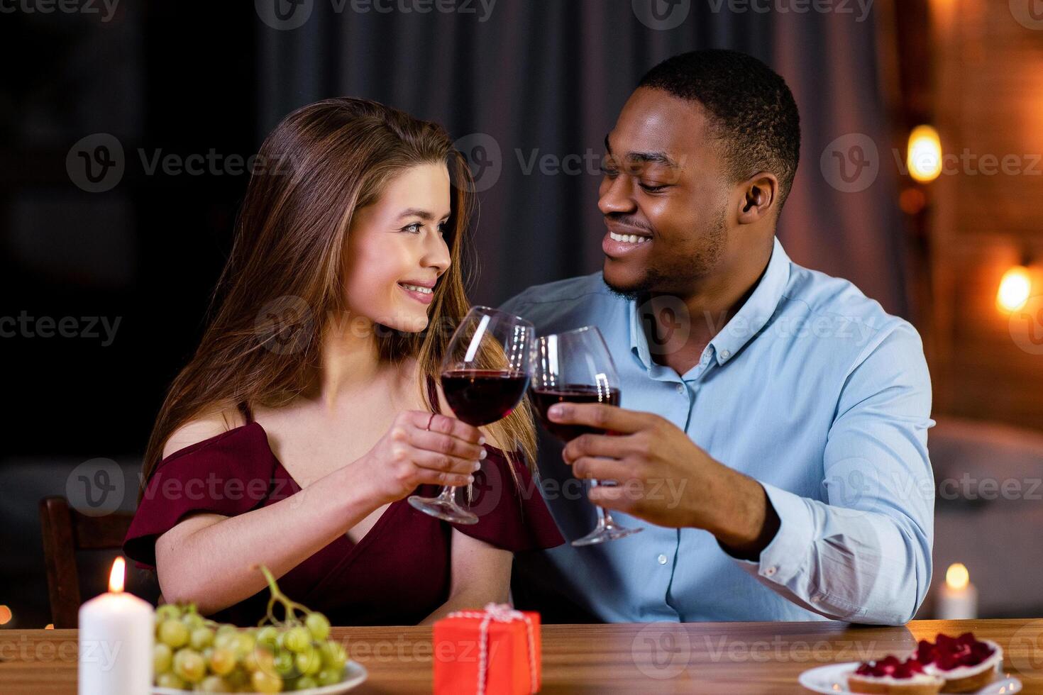 Romantic Dinner Concept. Happy Multiracial Couple Black Man And White Woman Celebrating Valentine's Day Together, Drinking Red Wine And Clinking Glasses While Dining In Restaurant, Free Space photo