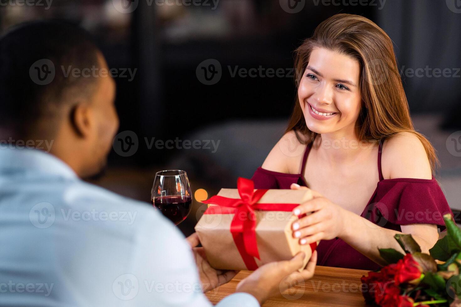 Happy Anniversary. Romantic Man Giving Wrapped Gift Box To His Girlfriend At Resraurant, Greeting With Valentine's Day, Loving Interracial Couple Dining In Elegant Atmosphere, Selective Focus photo