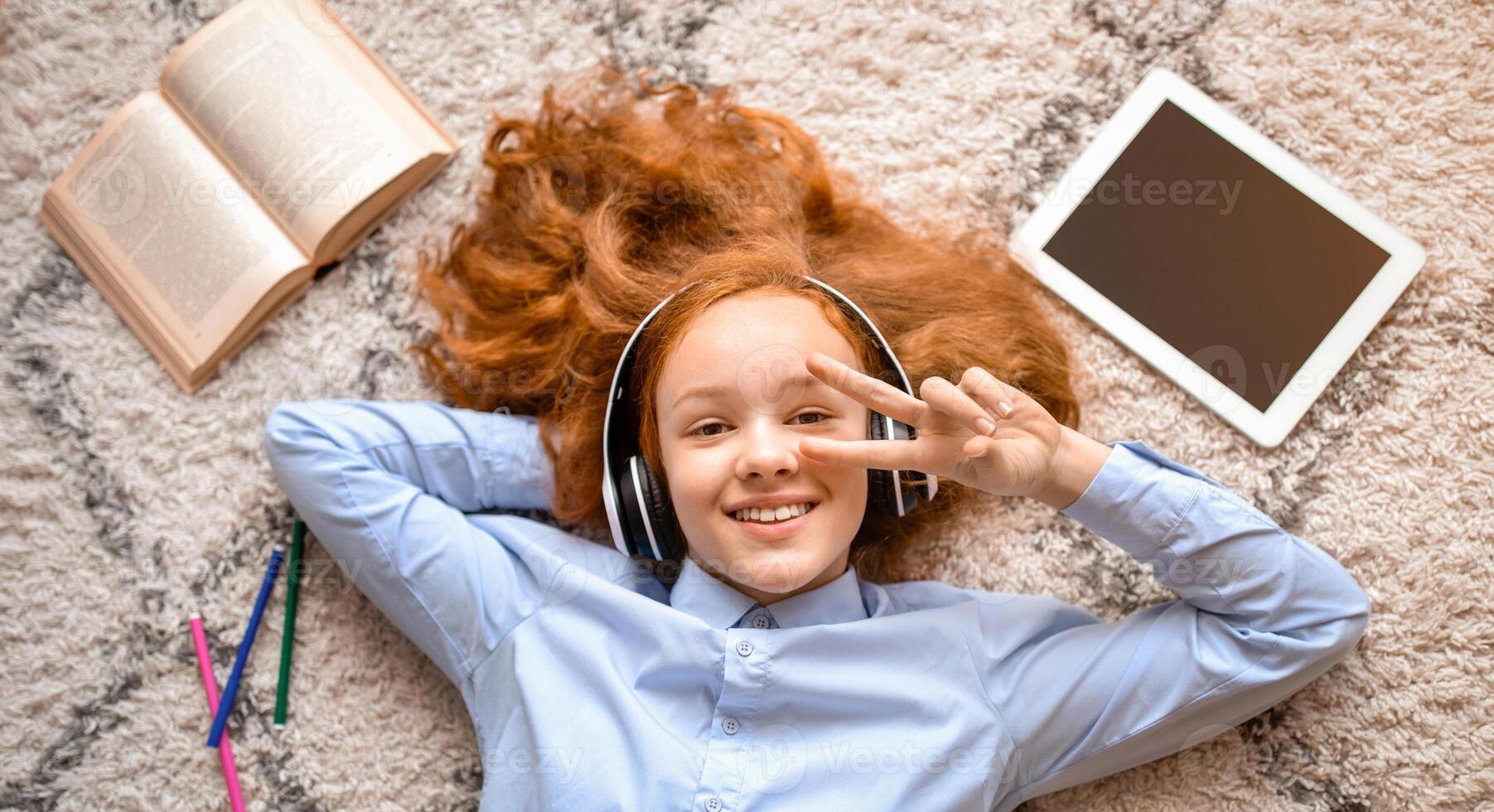 Top above high angle view of positive smiling red-haired teenage girl listening to music, enjoying her favourite song wearing wireless headphones, lying on floor carpet, showing v sign photo