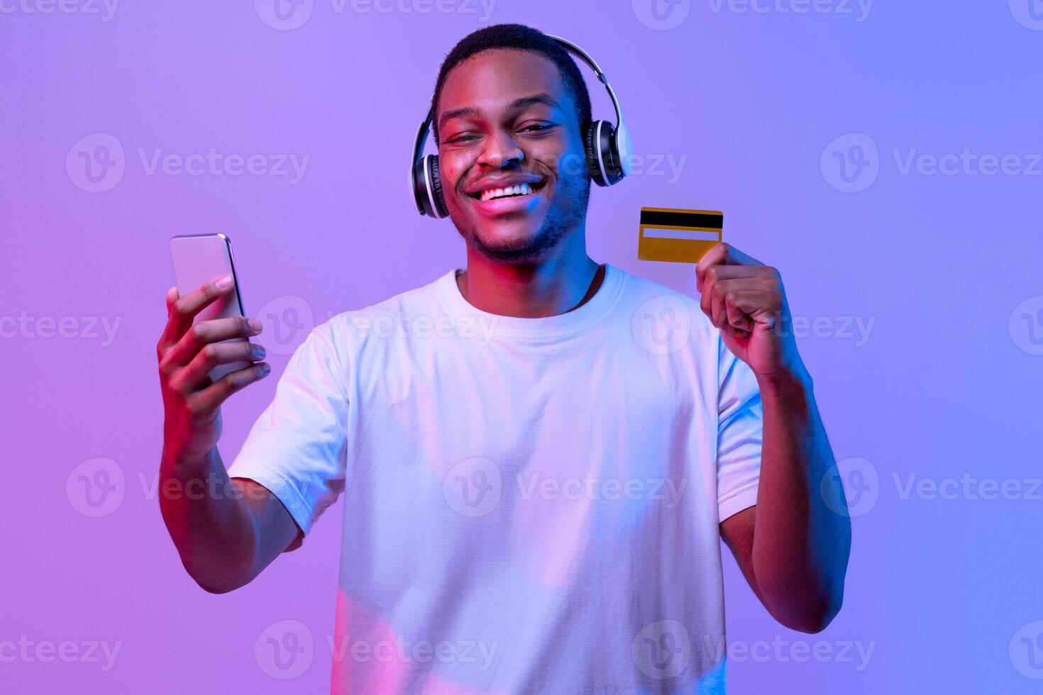 Streaming Music Subscription. Happy black guy in wireless headphones holding smartphone and credit card, paying for audio services online, standing under neon lighting over purple studio background photo