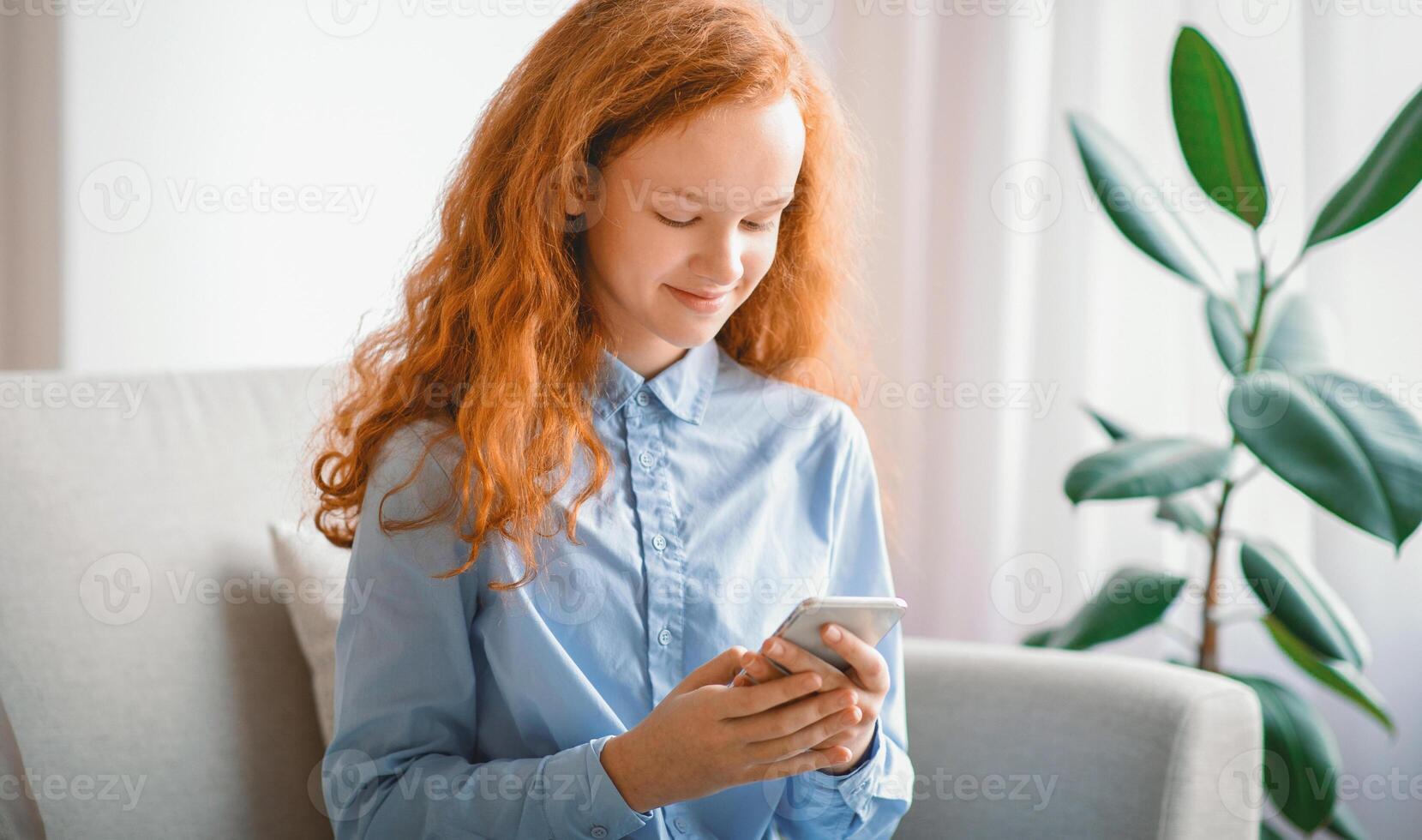 Digital Natives Concept. Positive smiling red-haired teenage kid using her cellphone, watching online, sitting on the couch in living room or bedroom at home. Girl scrolling social media feed photo