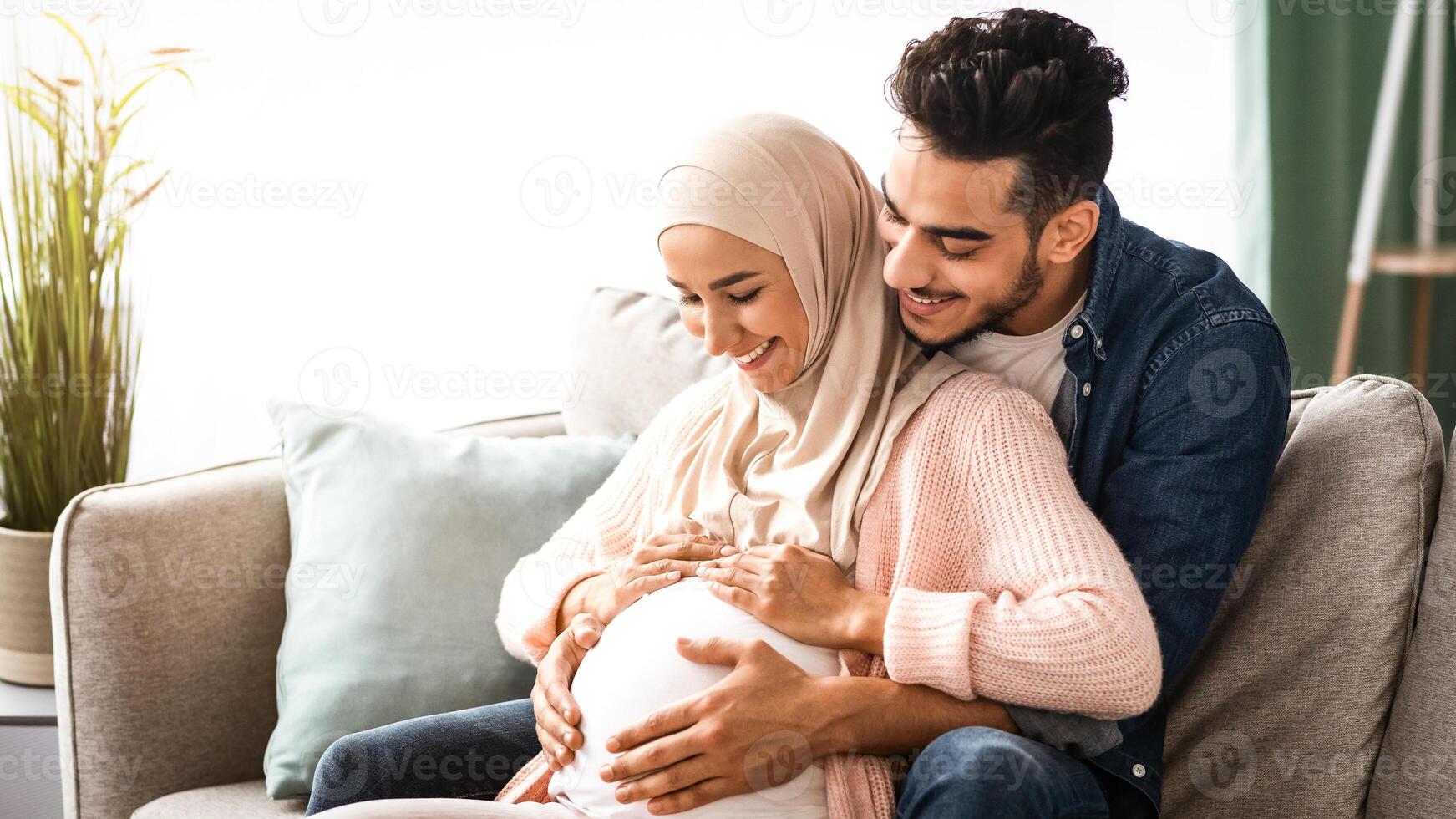 Happy islamic couple expecting baby, hugging on couch at home, cheerful muslim spouses relaxing together on sofa in living room, tenderly touching pregnant belly and smiling, copy space photo