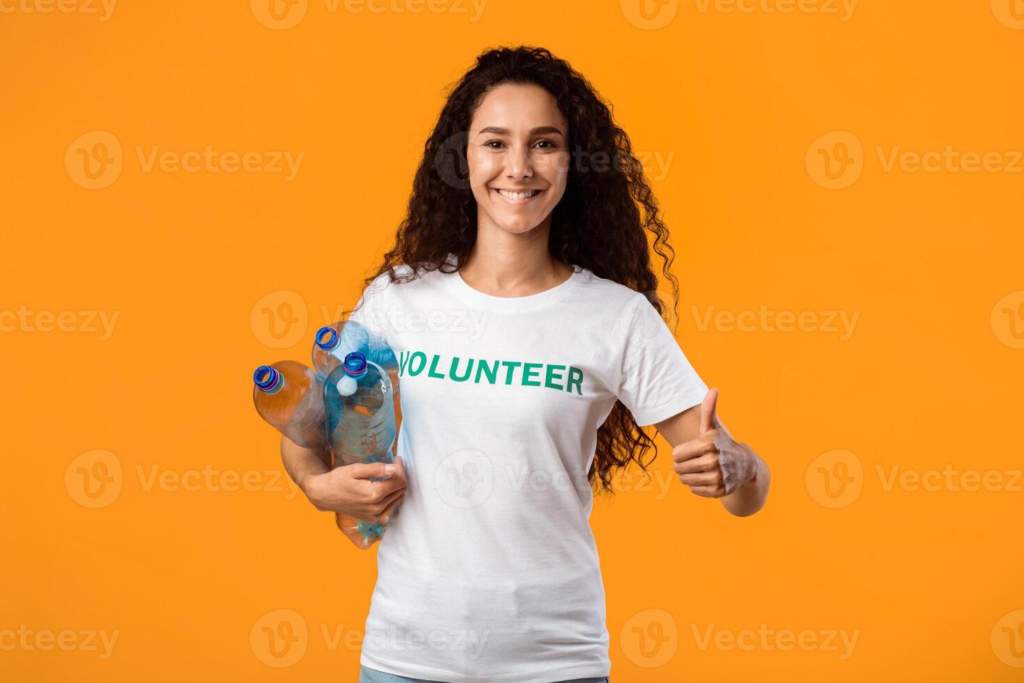 Happy Volunteer Lady Holding Plastic Bottles And Gesturing Thumbs Up To Camera Standing Over Yellow Studio Background. Recycle Your Waste, Keep Environment Clean photo