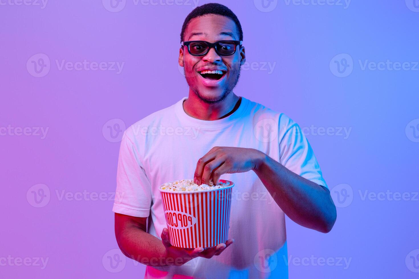 Portrait Of Emotional Black Guy In 3D Glasses With Bucket Of Popcorn, Cheerful African Man Looking At Camera With Excitement, Enjoying Good Movie, Standing Under Neon light Over Purple Background photo
