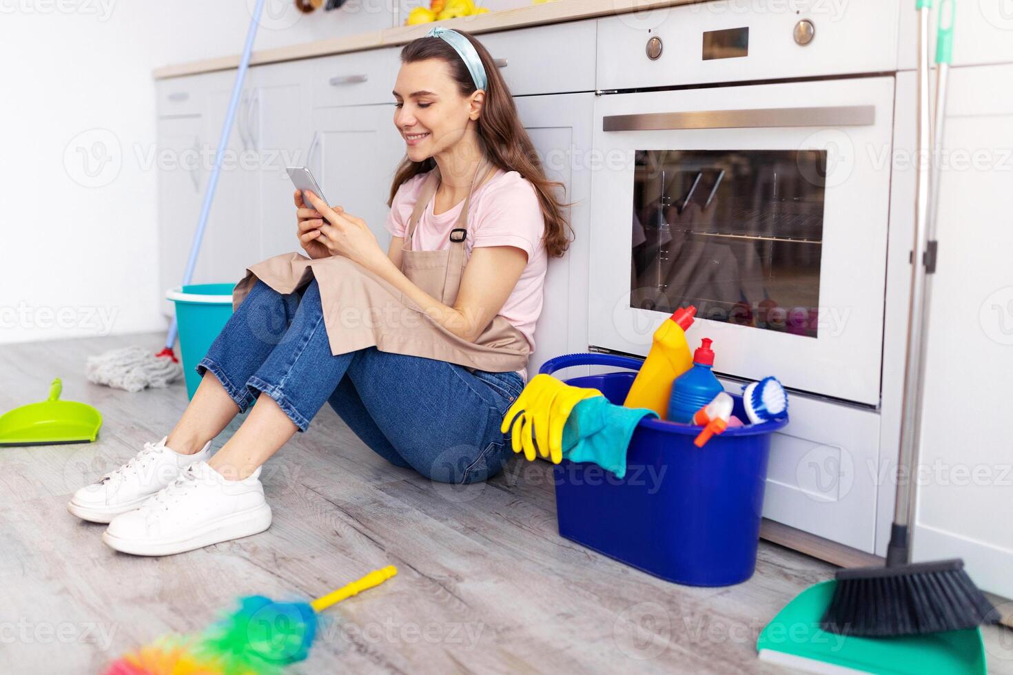 Taking break from house cleaning. Cheerful young housewife resting from domestic chores, using smartphone on floor in kitchen. Positive maid with cleaning supplies browsing web on movile device photo