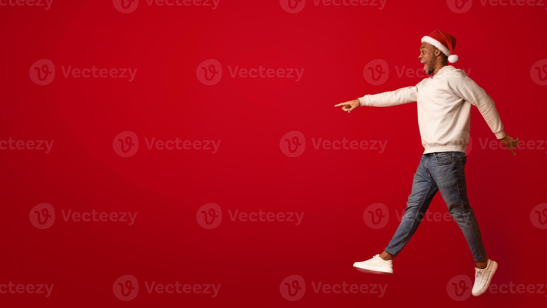 Check This. Excited Black Guy In Santa Hat Pointing Aside At Copy Space While Jumping On Orange Background, Showing Free Place For Your Xmas Offer Or Advertisement, Full Length Shot, Side View photo
