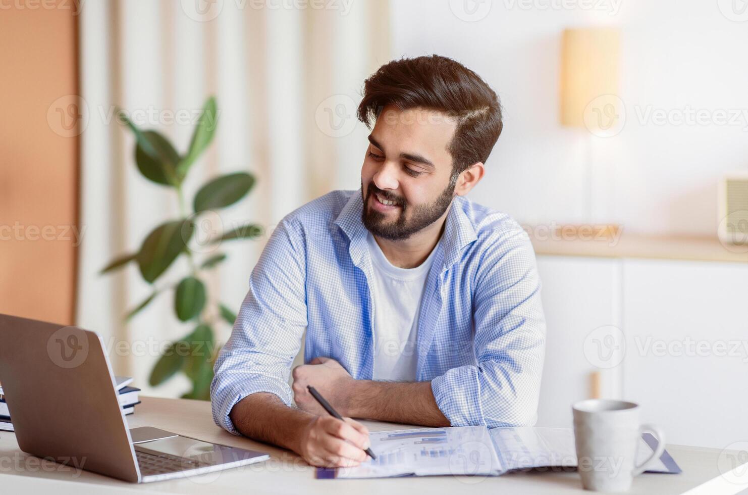 Successful Eastern Entrepreneur Working With Documents And Laptop At Home Office, Smiling Arab Businessman Looking At Computer Screen, Checking Financial Reports And Taking Notes, Free Space photo