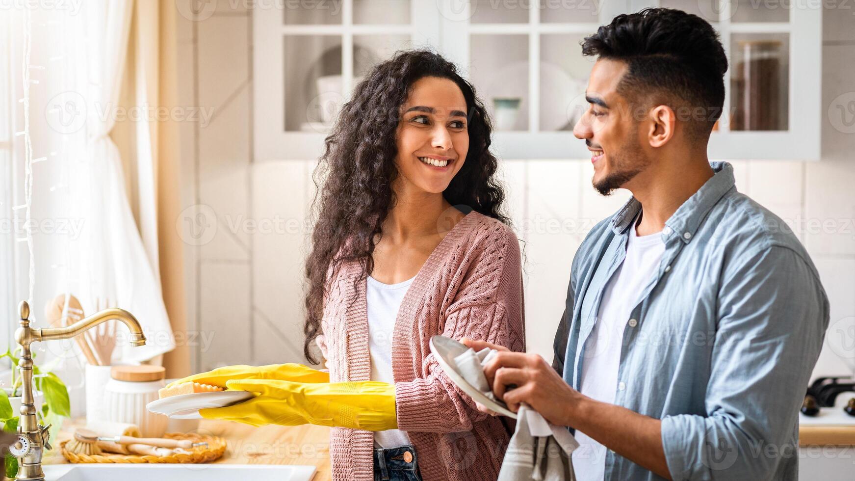 Happy Young Arab Spouses Washing Dishes In Kitchen Together, Loving Middle Eastern Couple Sharing Domestic Chores, Millennial Man And Woman Cleaning Home And Smiling To Each Other, Copy Space photo