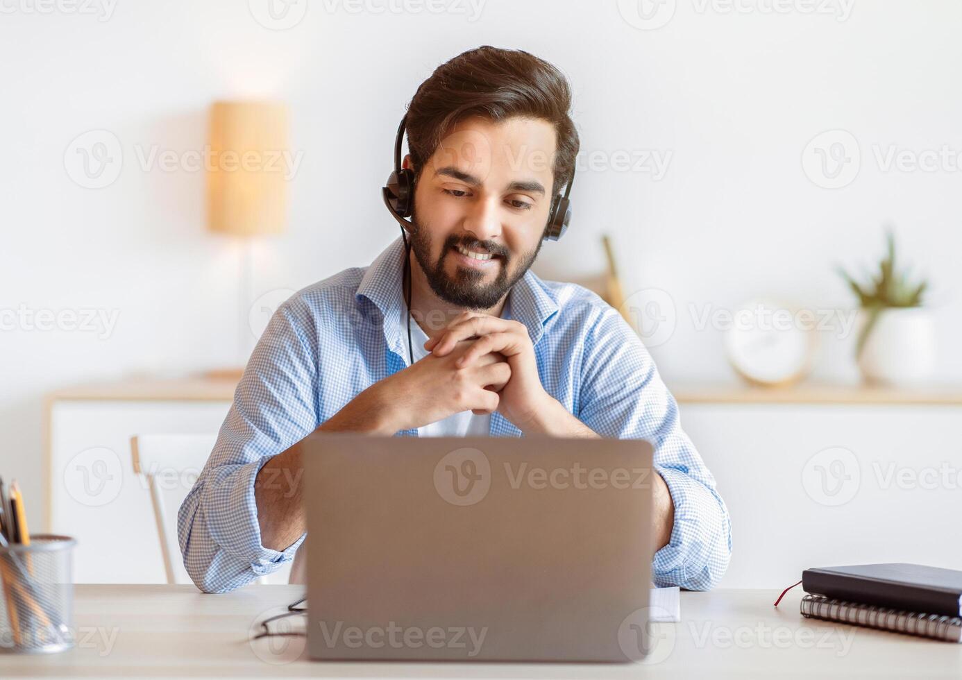 Portrait of smiling arab guy in headset watching webinar on laptop, young eastern man sitting at desk and looking at computer screen, enjoying online education and distance learning, free space photo
