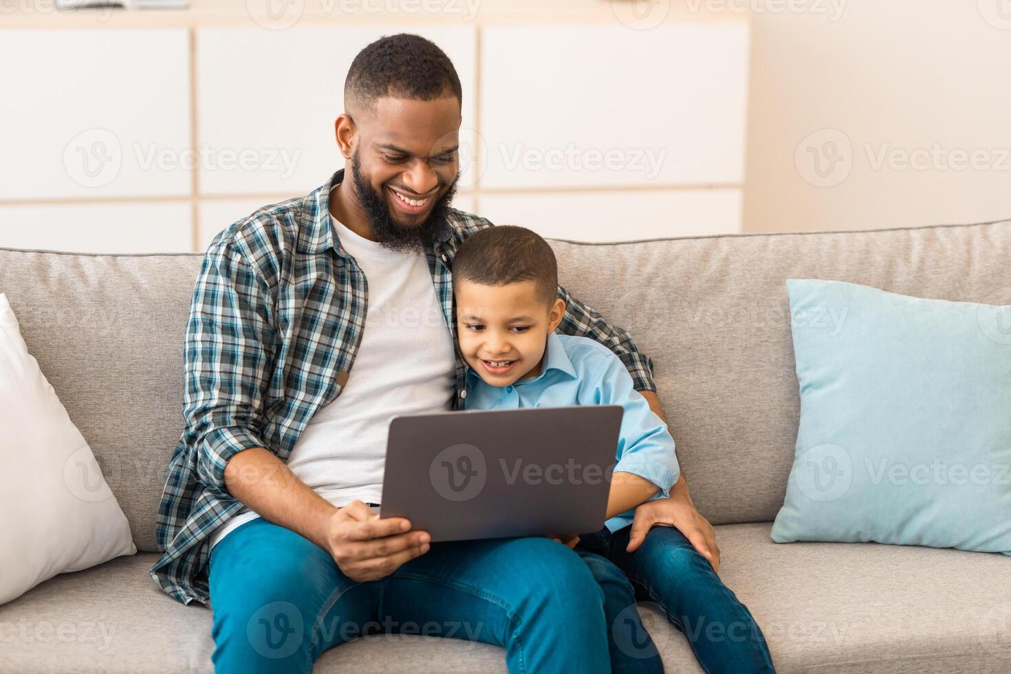 African Daddy And Son Watching Cartoons Online On Laptop Computer Sitting On Sofa Together At Home. Weekend With Father. Family Lifestyle And Gadgets Concept photo