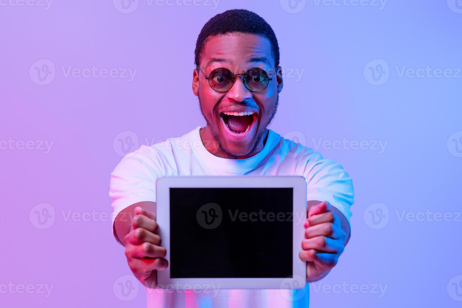Excited African American Guy Showing Digitral Tablet With Black Screen At Camera, Demonstrating Free Copy Space For Your Website Or App Mockup Design, Standing In Neon Light Over Purple Background photo