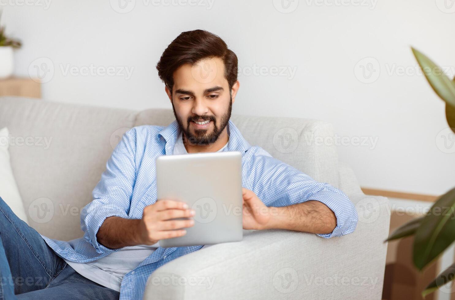 Handsome Eastern Man Browsing Internet On Digital Tablet While Relaxing At Home. Young Smiling Arab Guy Sitting On Couch In Living Room Using Tab Computer, Shopping Online Or Surfing Web, Free Space photo