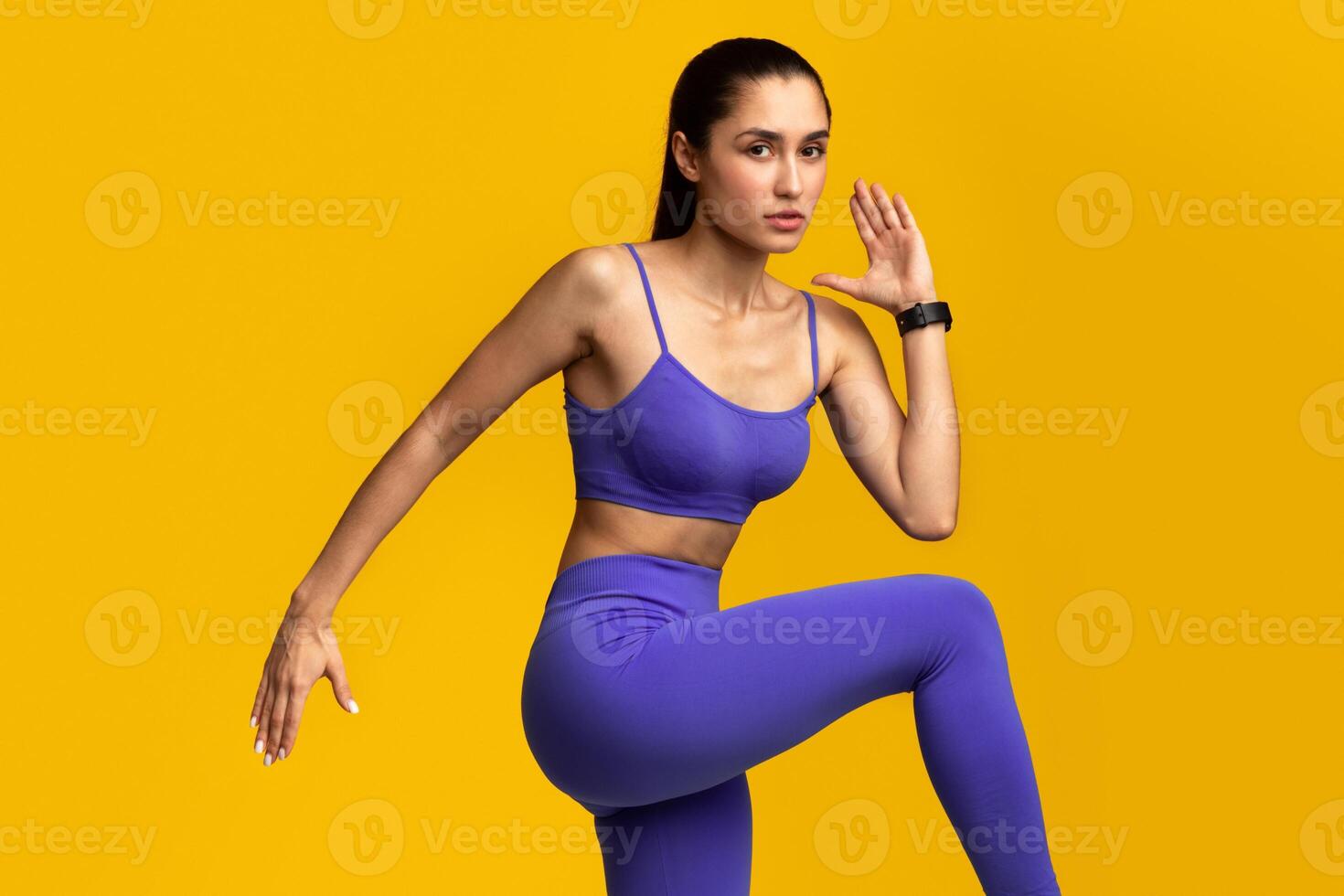 Fitness And Sport Concept. Portrait Of Confident Fit Young Lady Wearing Activity Tracker Running, Lifting Leg Up, Exercising Isolated On Orange Studio Background, Looking At Camera. Energy And Workout photo