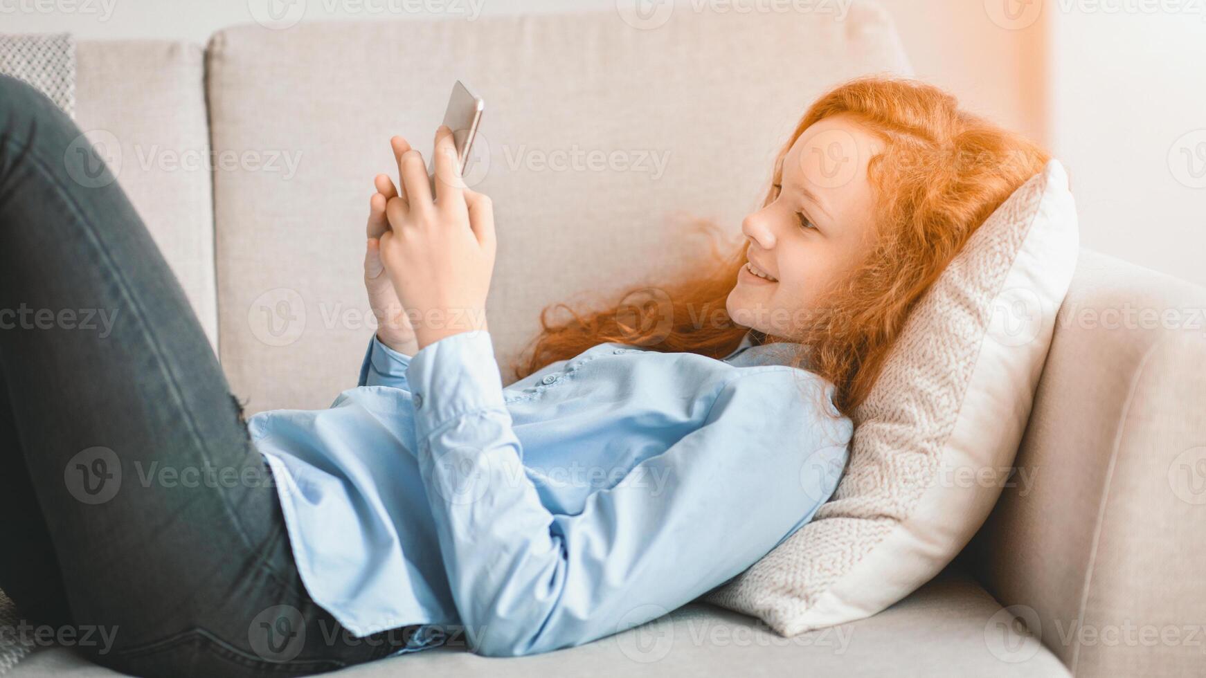 Digital Natives. Portrait of smiling red-haired teen girl using her cellphone, watching online, resting and lying on the couch in living room at home. Child having fun spending time alone photo