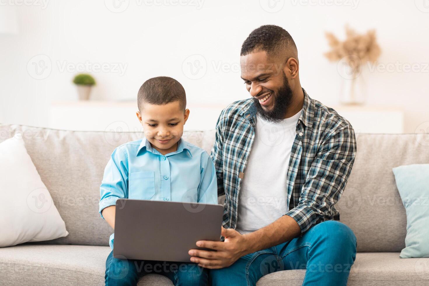 African American Boy And His Father Using Laptop Together, Watching Cartoons Online On Computer Sitting On Sofa At Home. Young Black Dad Bonding With Son Browsing Internet On Gadget Indoor photo