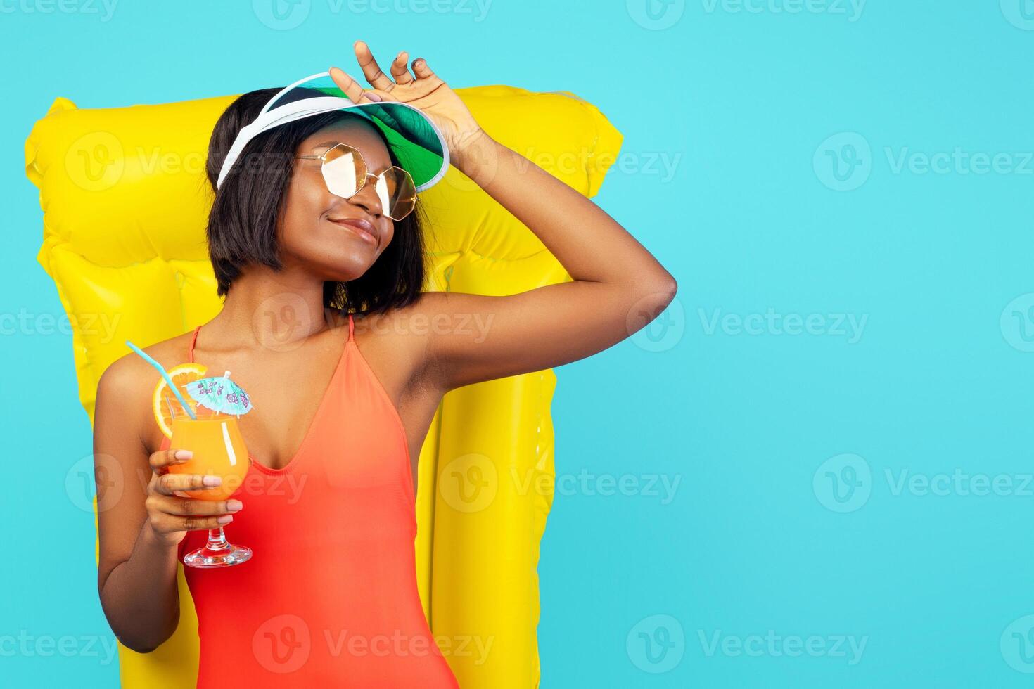 Pretty black lady in swimsuit lying on inflatable lilo and enjoying tropical cocktail over blue studio background. Beautiful African American woman relaxing on pool float, having summer chill photo