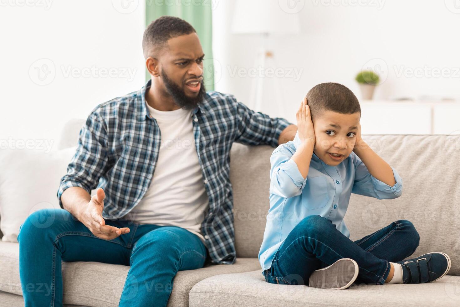 Irritated Black Father Shouting At Unhappy Little Son While He Covering Ears Sitting On Couch At Home. Domestic Violence Against Child, Bad Family Relationship Problem. Selective Focus photo