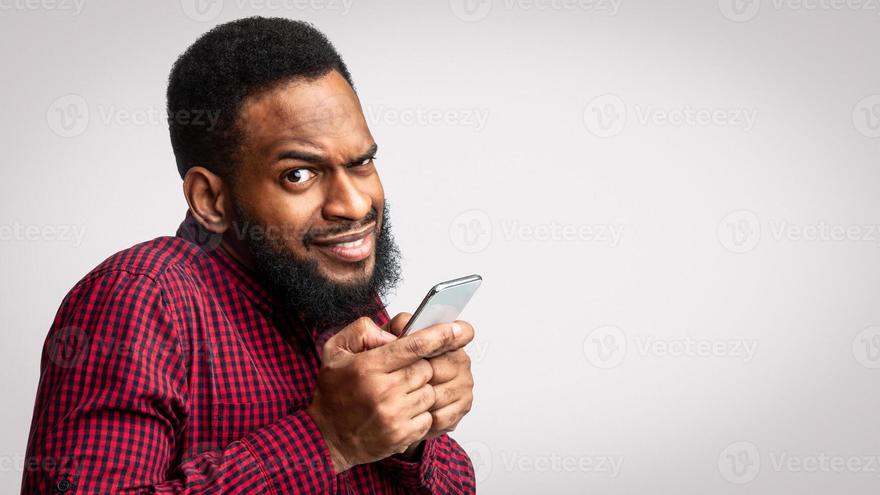 Phone Fraud. Suspicious Black Man Using Smartphone Looking At Camera Posing Over White Studio Background. Telephone Phreaking And Cellphone Scam Concept photo