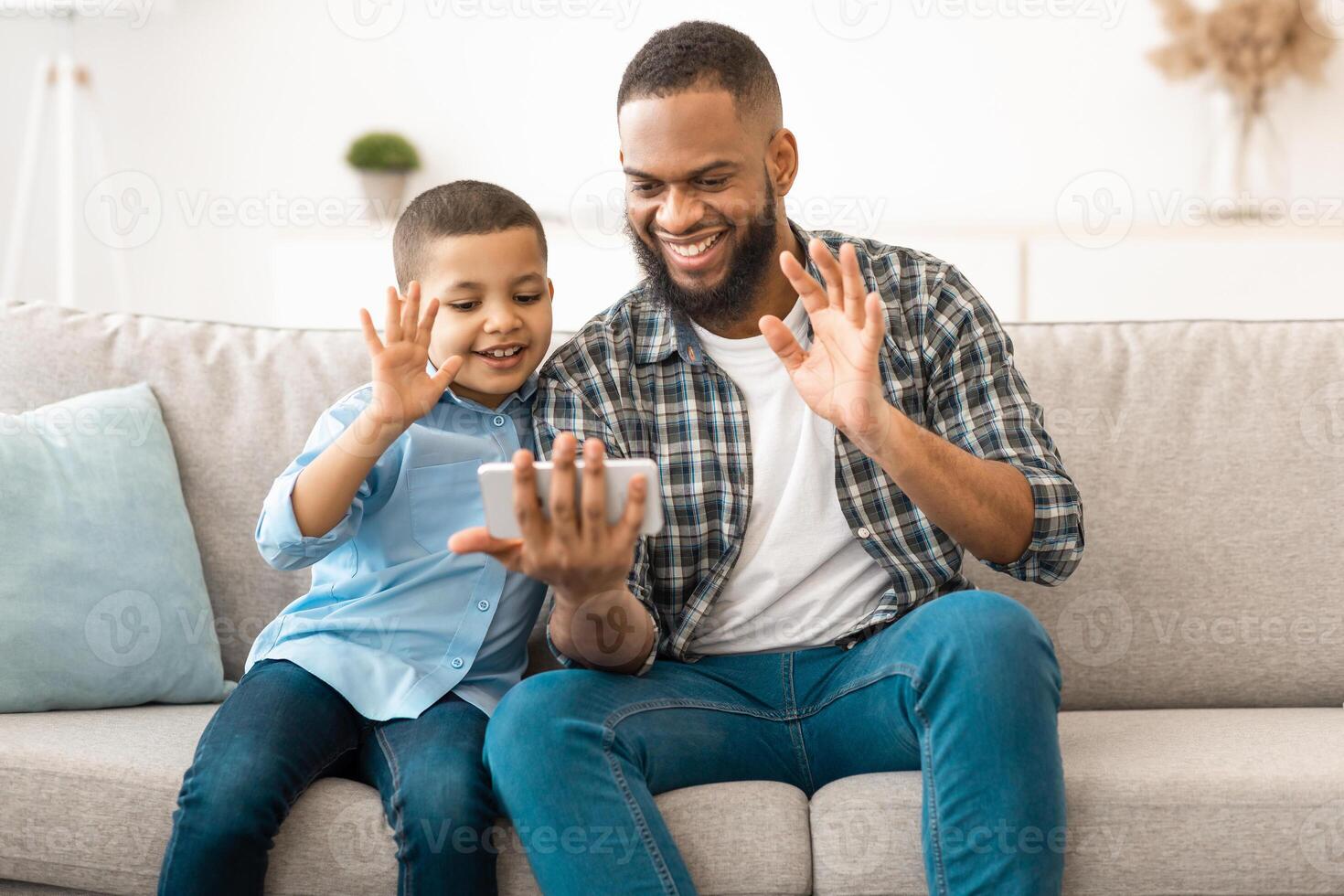 Call. African Father And Son Using Mobile Phone Calling Waving Hands To Smartphone Screen Sitting On Sofa At Home. Remote Online Communication Applications For Cellphone photo